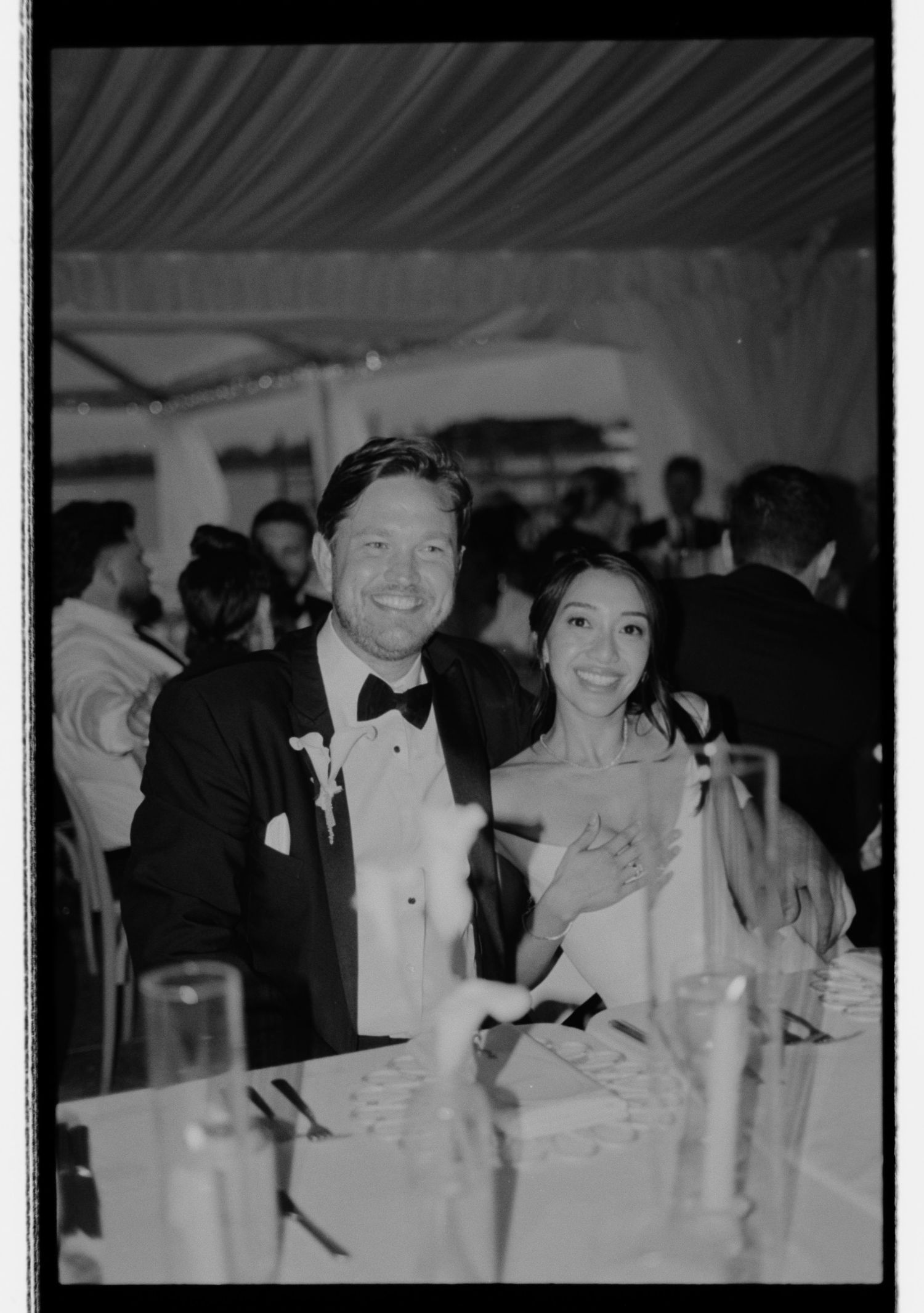 Black and white photo of a smiling couple in formal attire seated at a wedding reception dinner table.