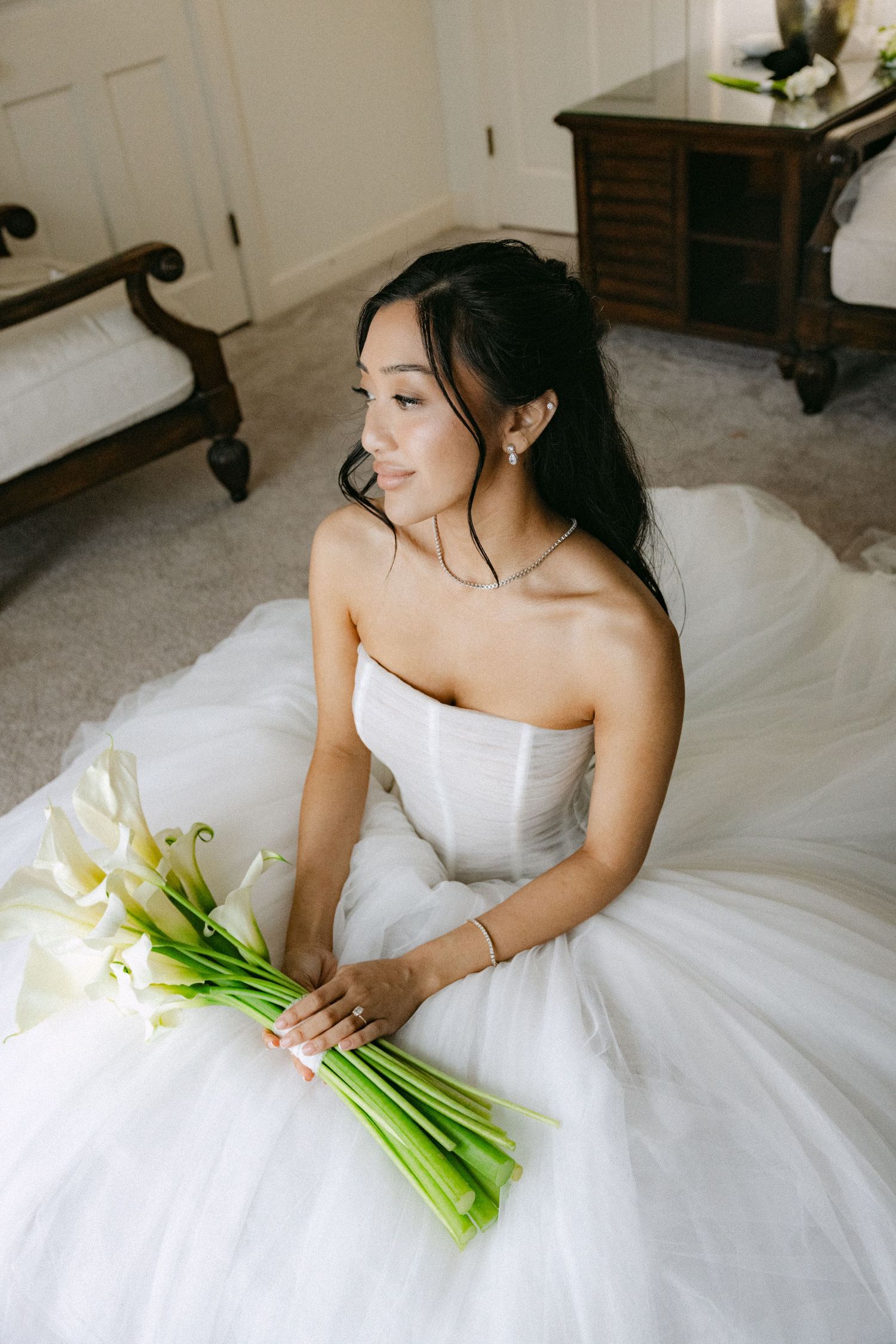 Bride in strapless white ballgown sits on floor holding calla lily bouquet, looking away in elegant bridal suite.