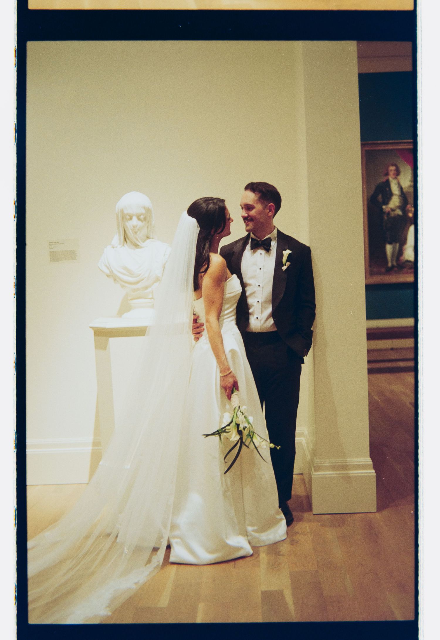 Bride and groom share a romantic moment in a museum gallery, with a white marble bust sculpture nearby.