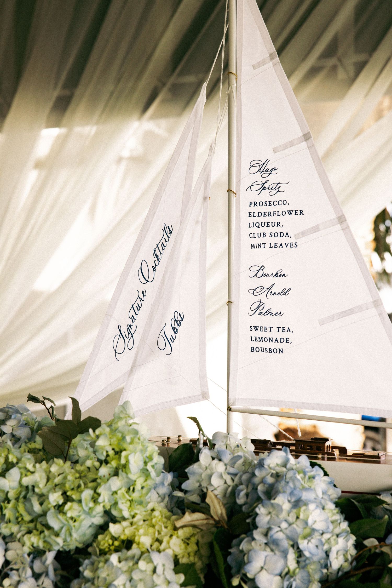Elegant nautical wedding decor featuring a model sailboat with calligraphy menus on sails, surrounded by blue hydrangeas.