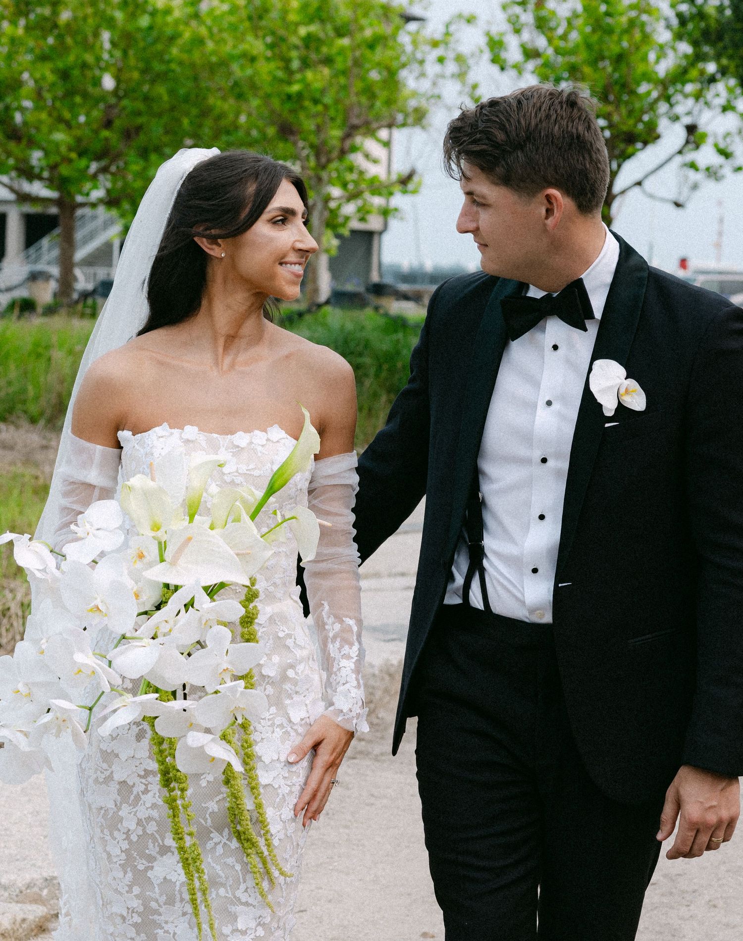 Elegant couple in wedding attire share candid moments while holding white orchid bouquet outdoors.