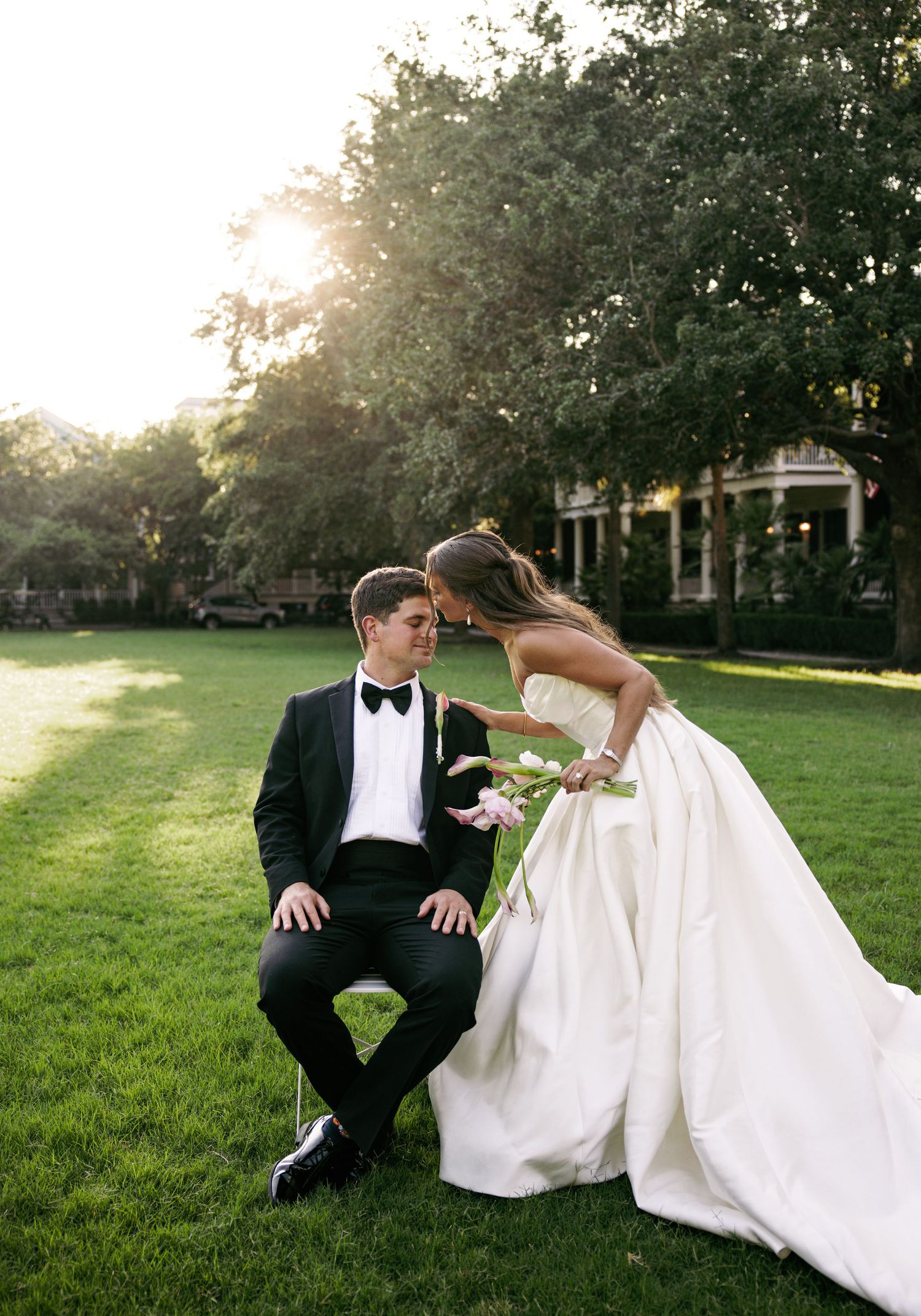 Bride in strapless white ballgown leans toward groom in black tuxedo seated on lush green lawn at golden hour.