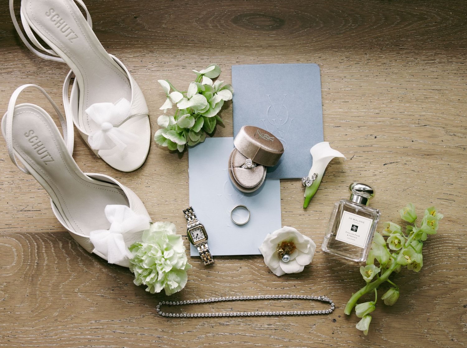 Bridal flat lay with white heels, watch, rings, perfume, necklace, and green floral accents on wood floor.