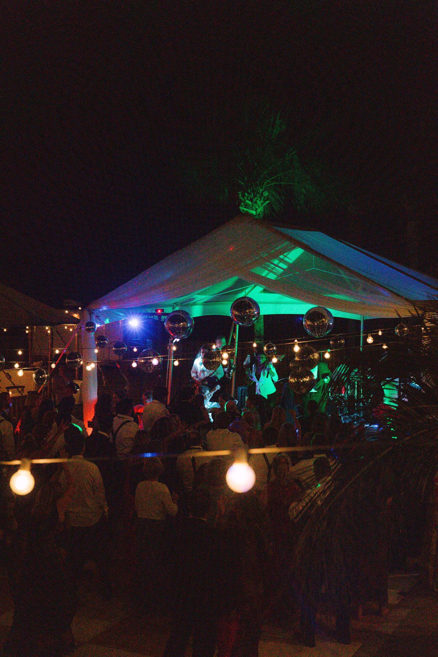 Live band performing under lit tent with colorful stage lights at outdoor wedding reception.