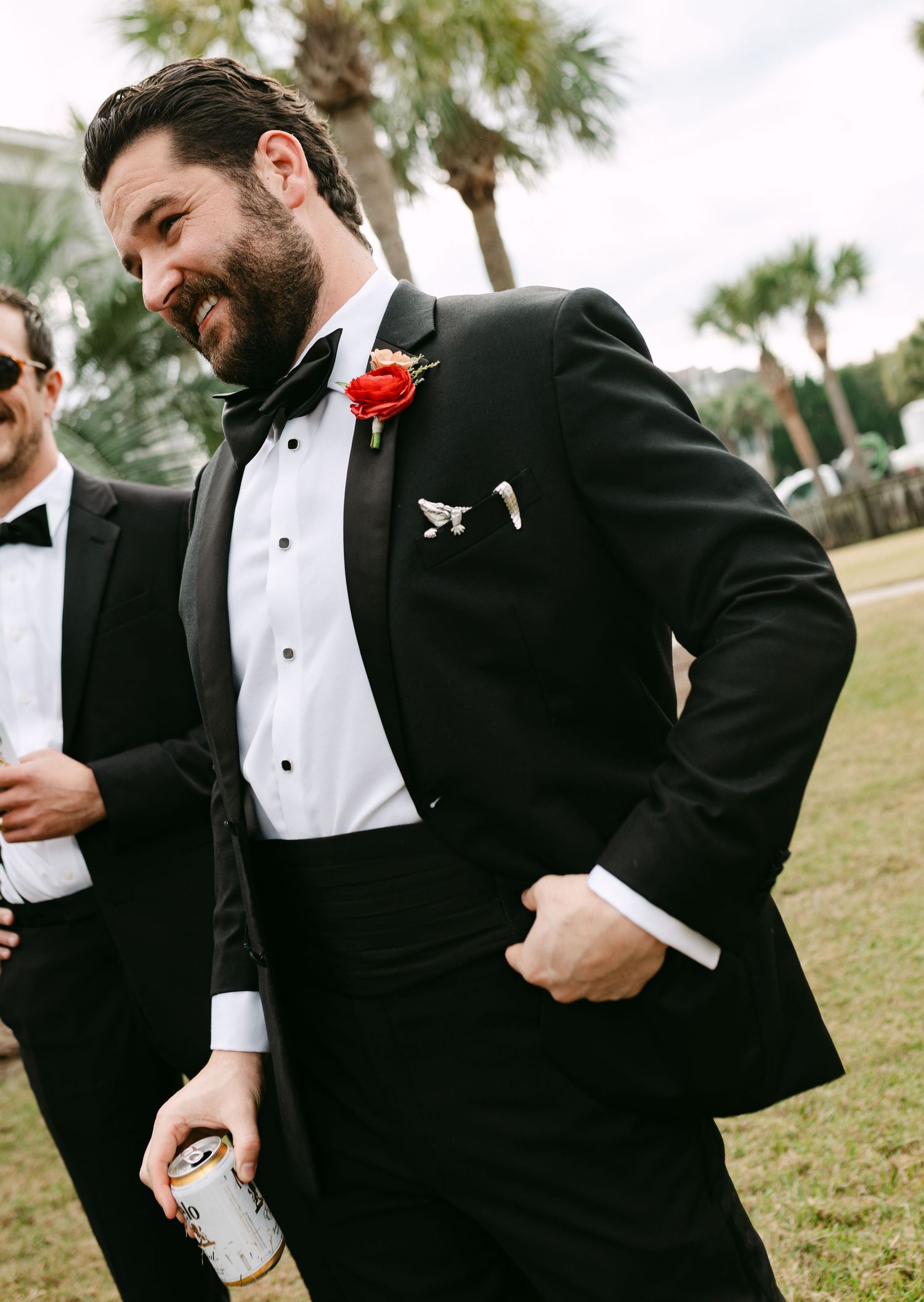 Bearded groom in black tuxedo with red rose boutonniere smiles while holding a beer can outdoors.