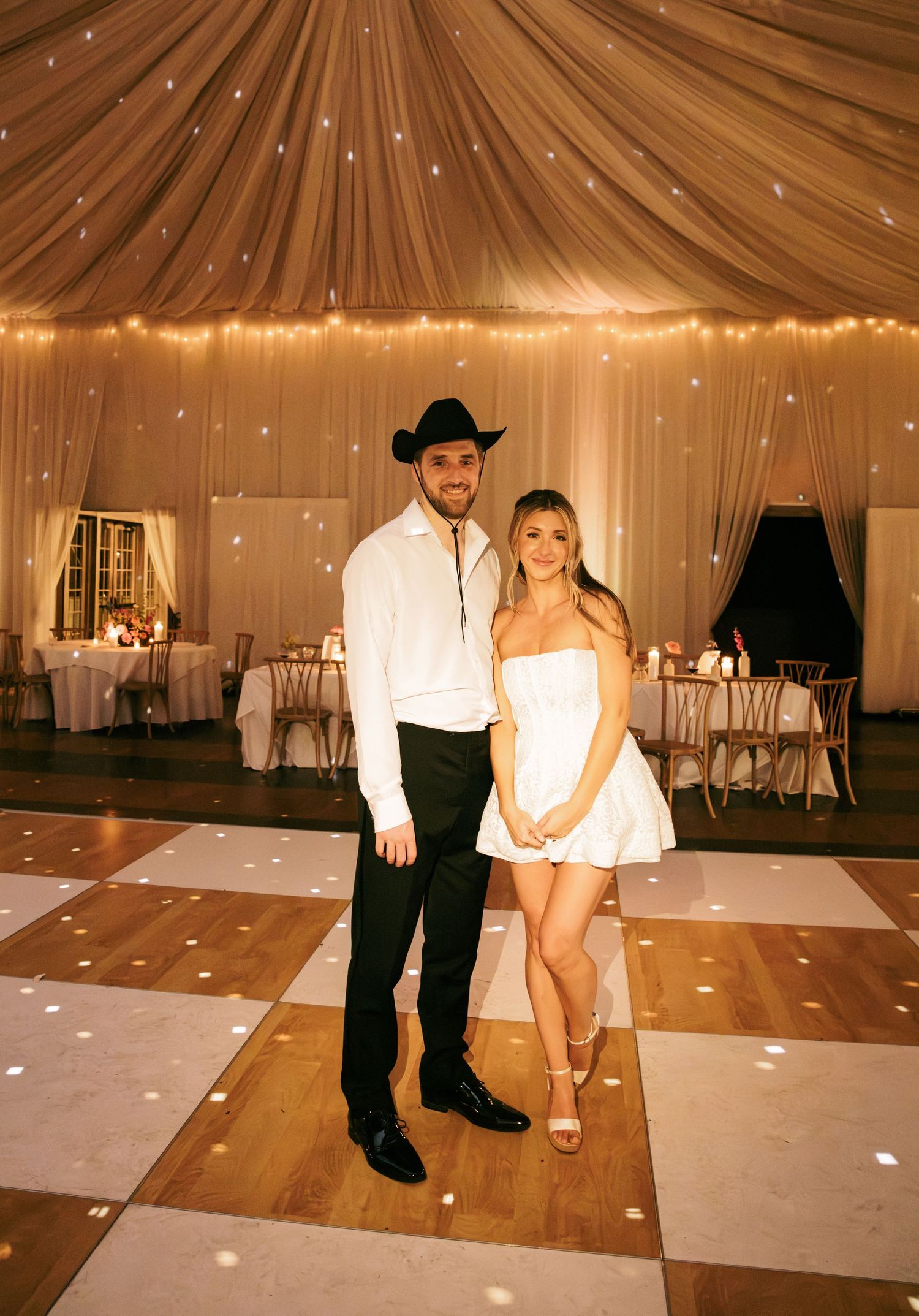 Couple posing on white dance floor in elegant tent venue with string lights, man in cowboy hat and woman in white mini dress.