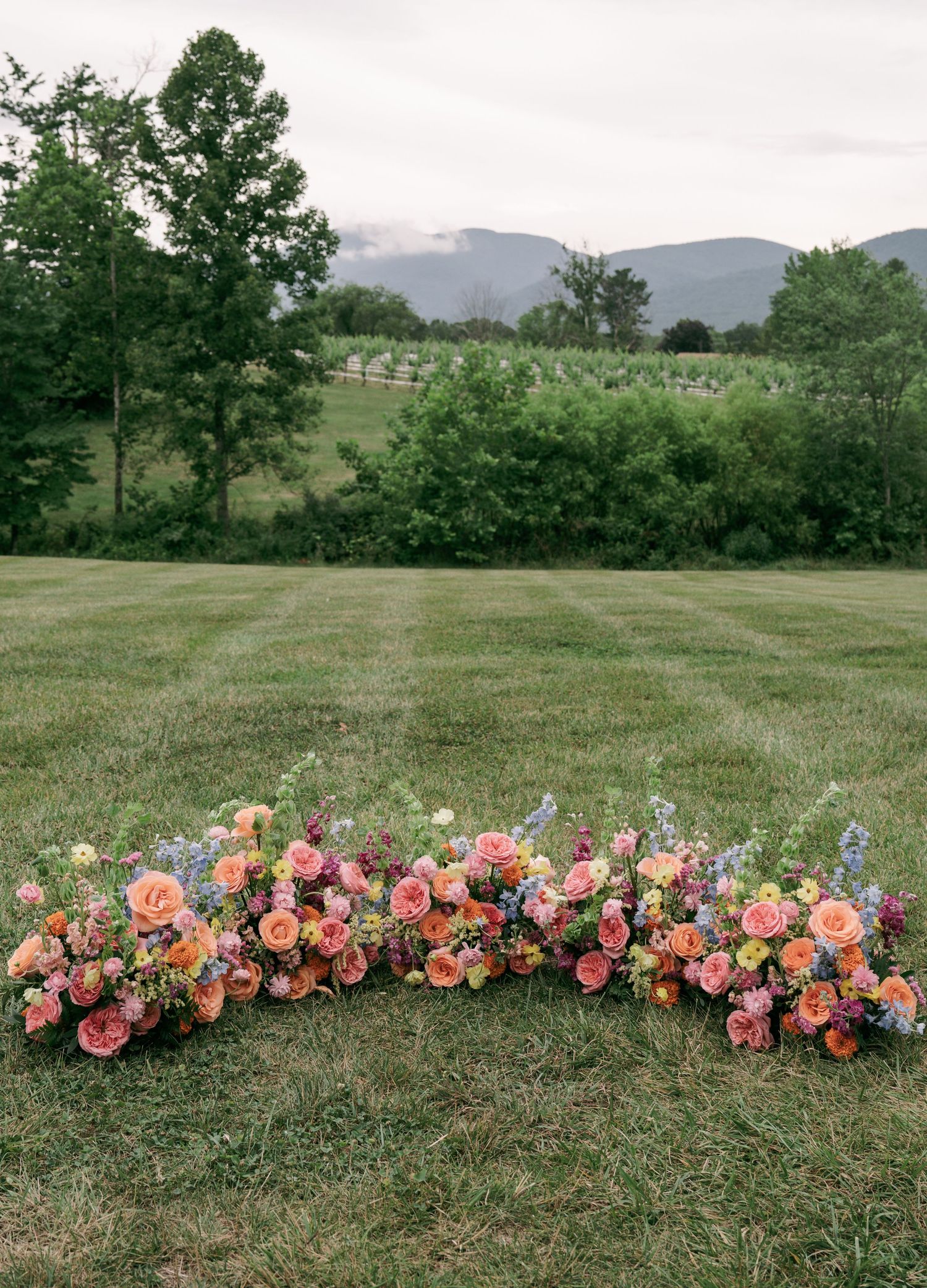 Colorful floral arrangement of pink, orange, and blue flowers on green lawn with mountain backdrop.