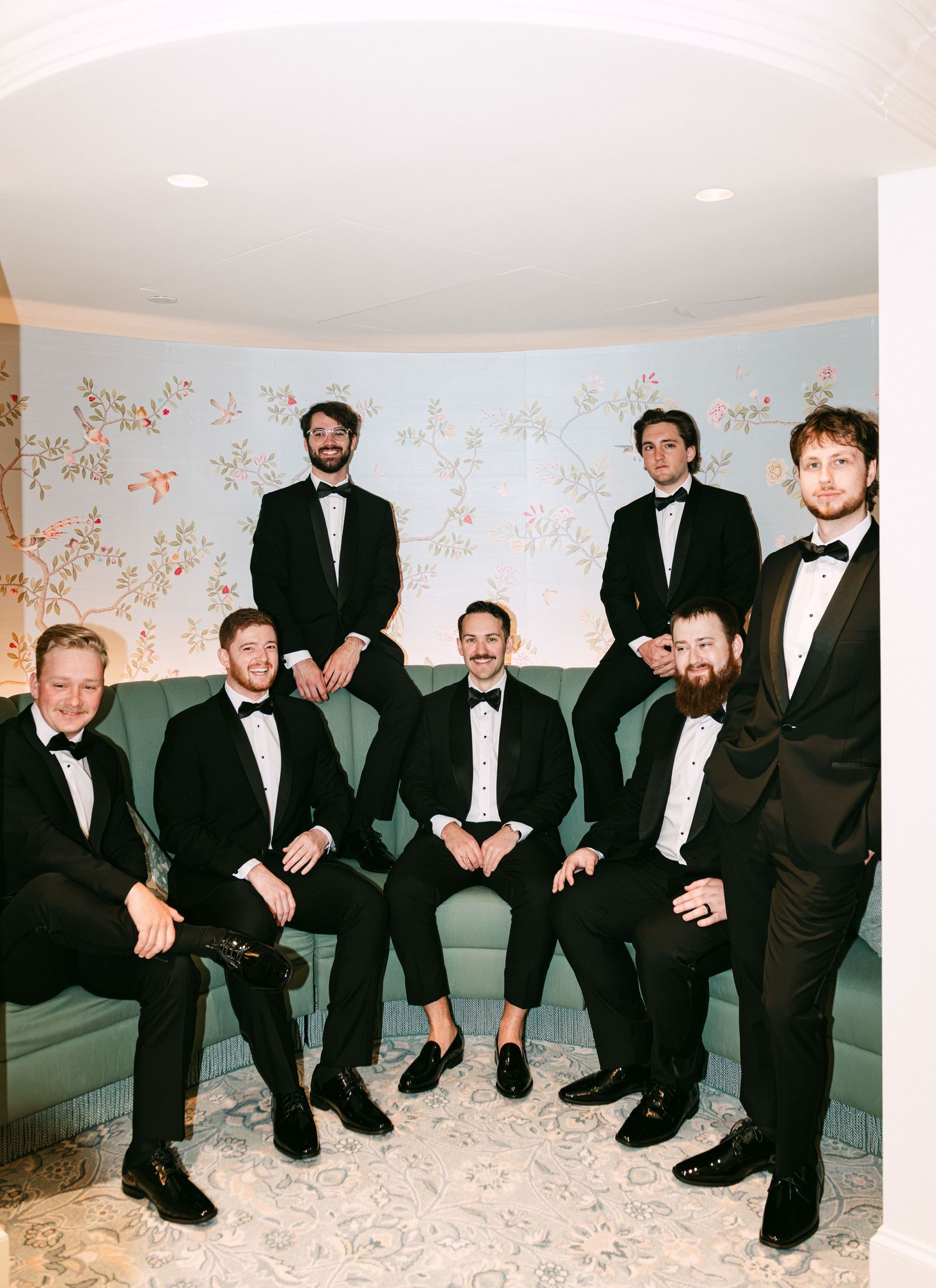 Groom and groomsmen in black tuxedos pose together in an elegant room with floral wallpaper.