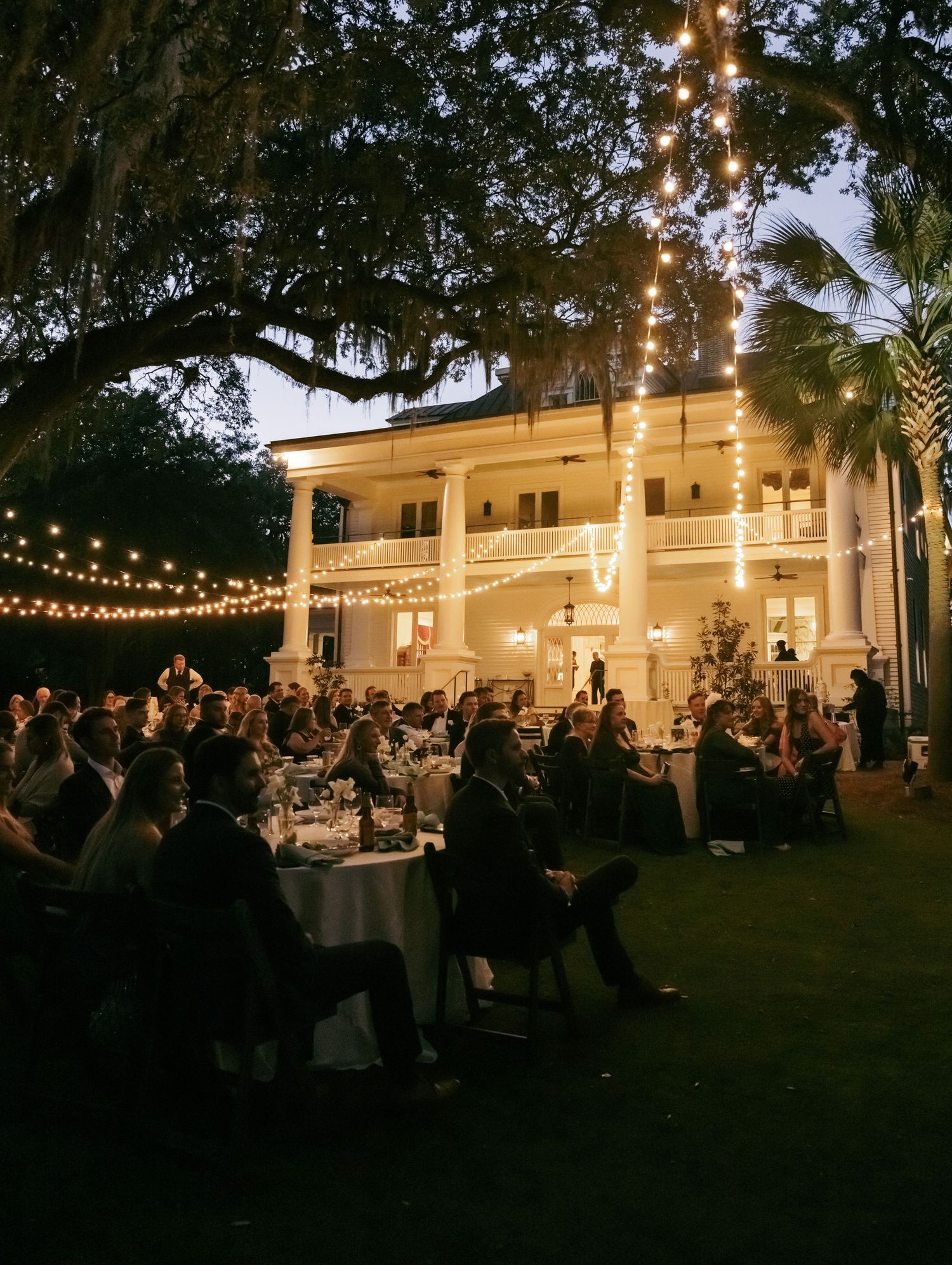 Elegant outdoor evening wedding reception with string lights on mansion lawn, guests dining under oak trees at dusk.