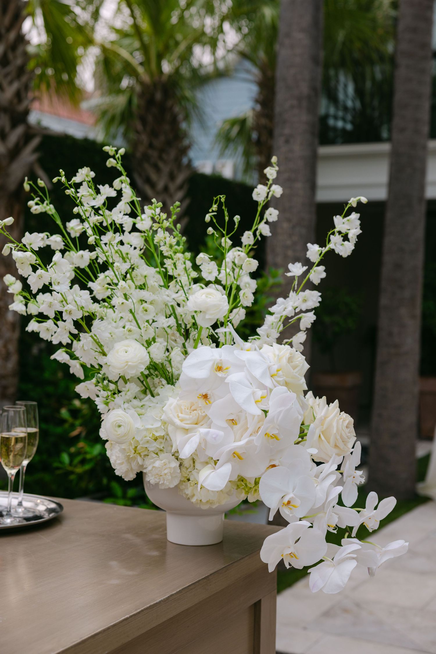 White floral arrangements with orchids and roses displayed on tables with palm trees in background.