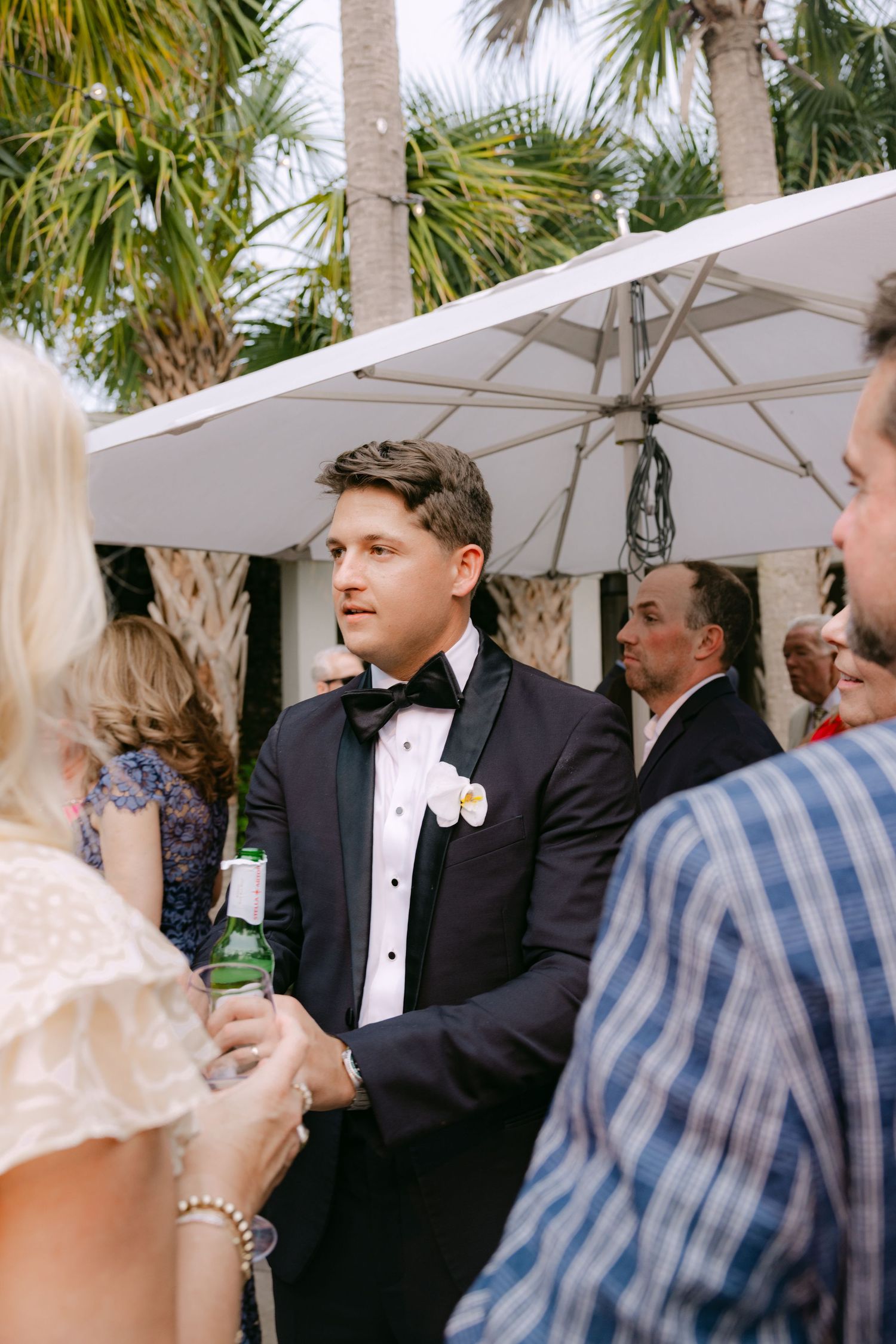 Outdoor wedding reception with guests mingling under white umbrellas and palm trees.