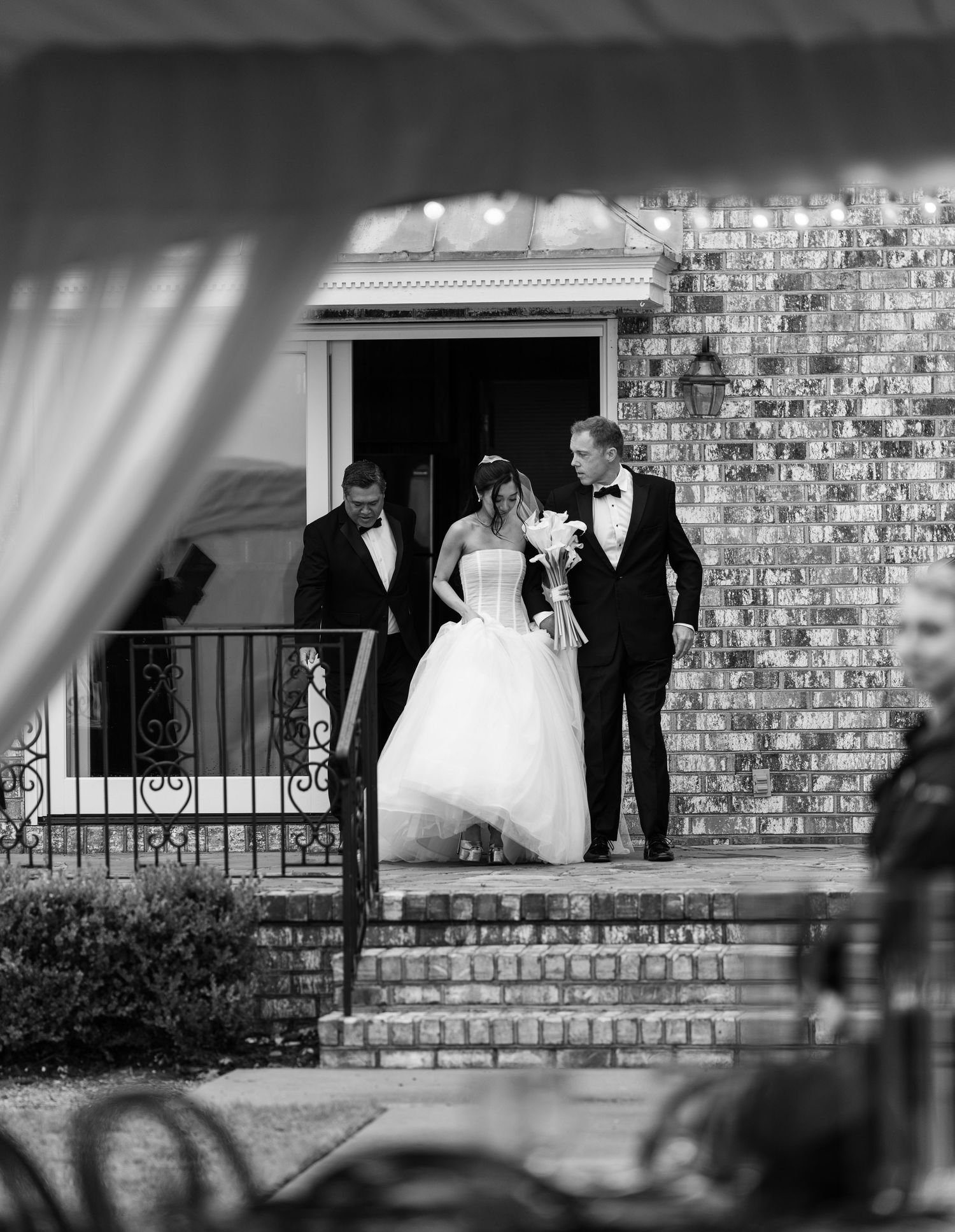 Black and white photo of a bride in a ball gown walking down church steps with two men in tuxedos.