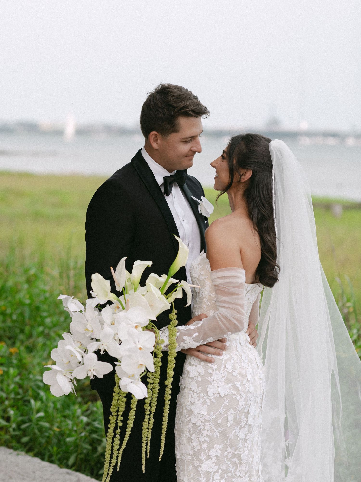 Elegant wedding couple embraces in an outdoor setting with white floral bouquet against a coastal backdrop.