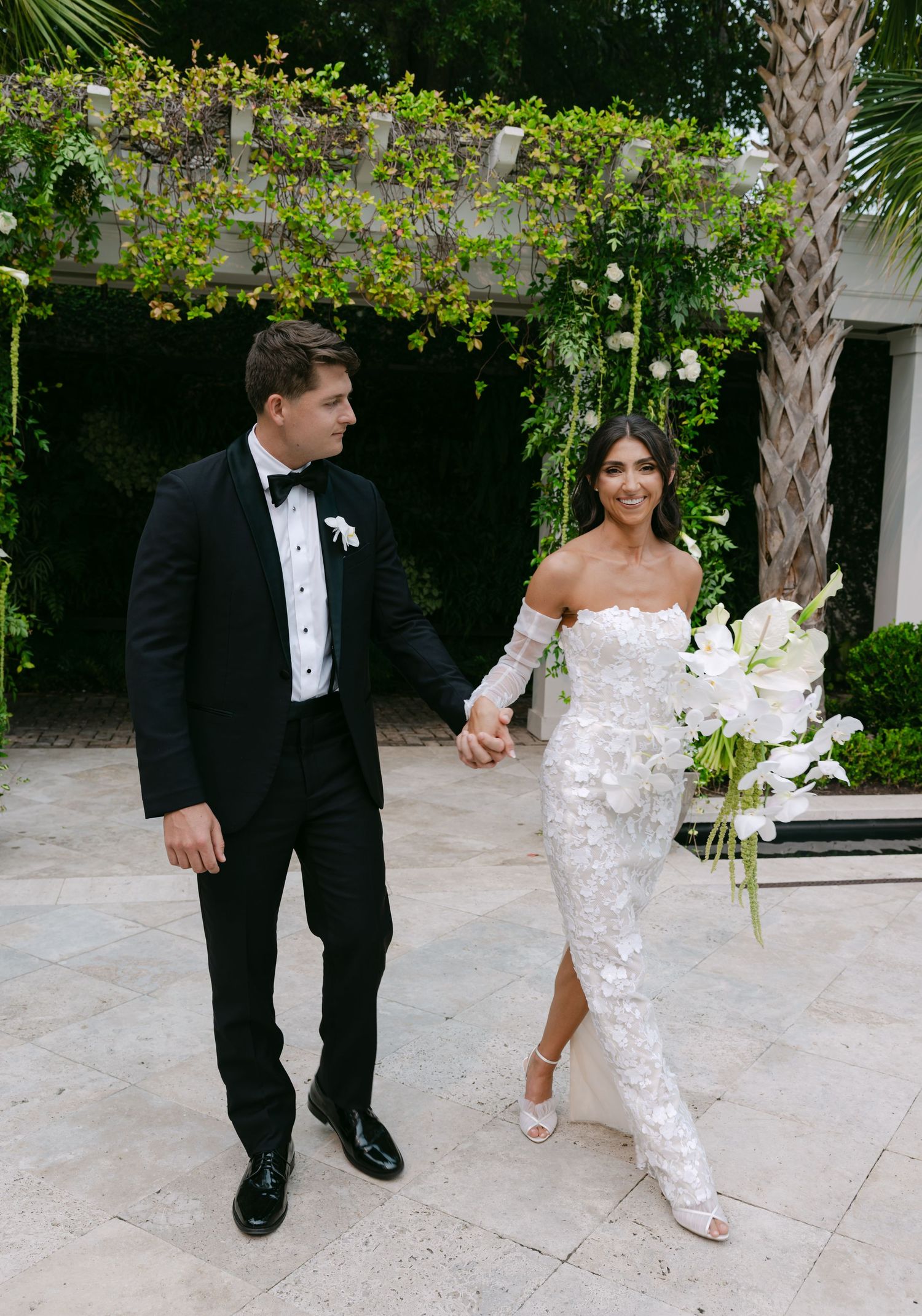 Elegant bride in lace gown and groom in tuxedo share a playful moment in a garden setting.