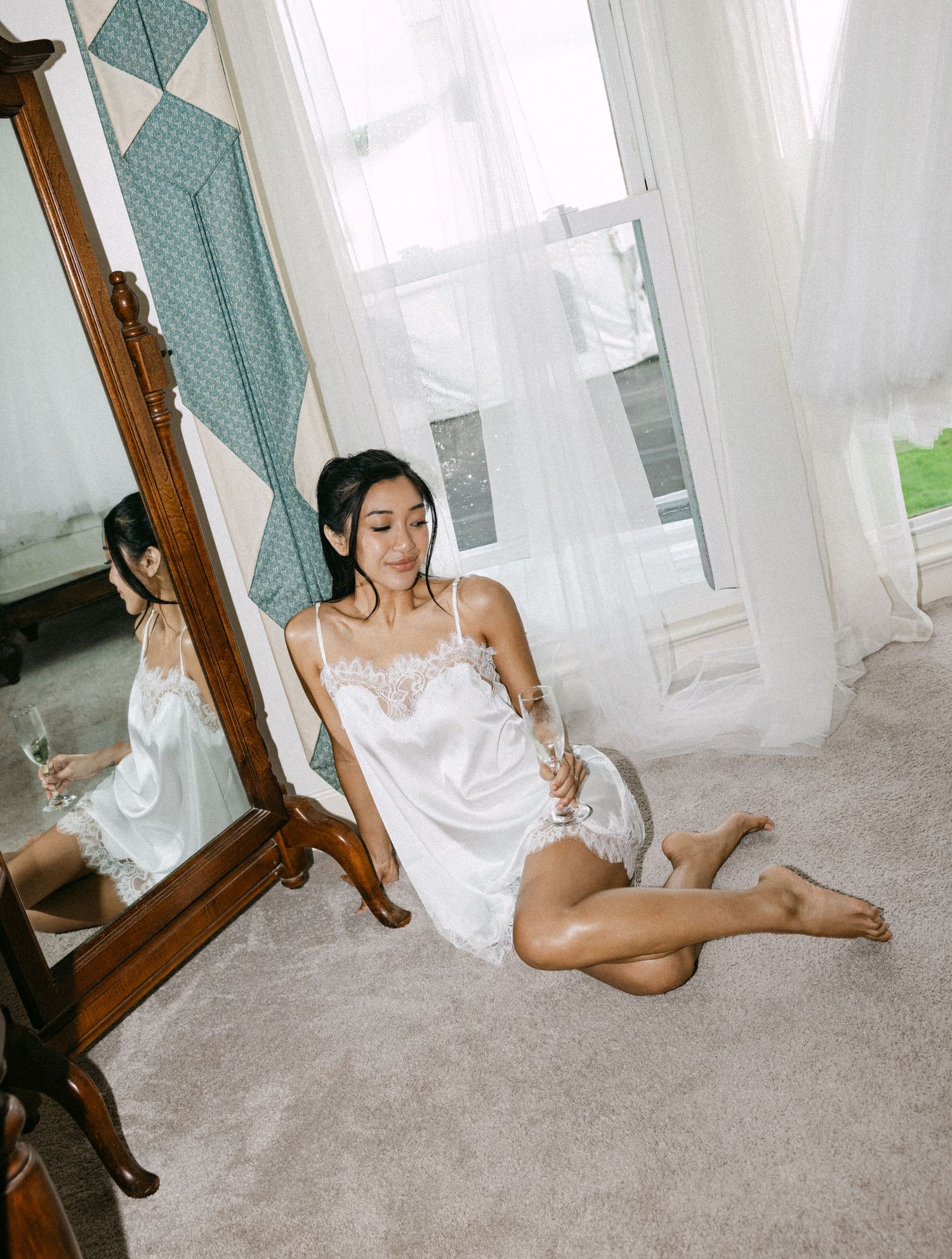 Woman in white lace slip dress sitting on carpet beside ornate mirror, holding champagne glass near sheer curtains.