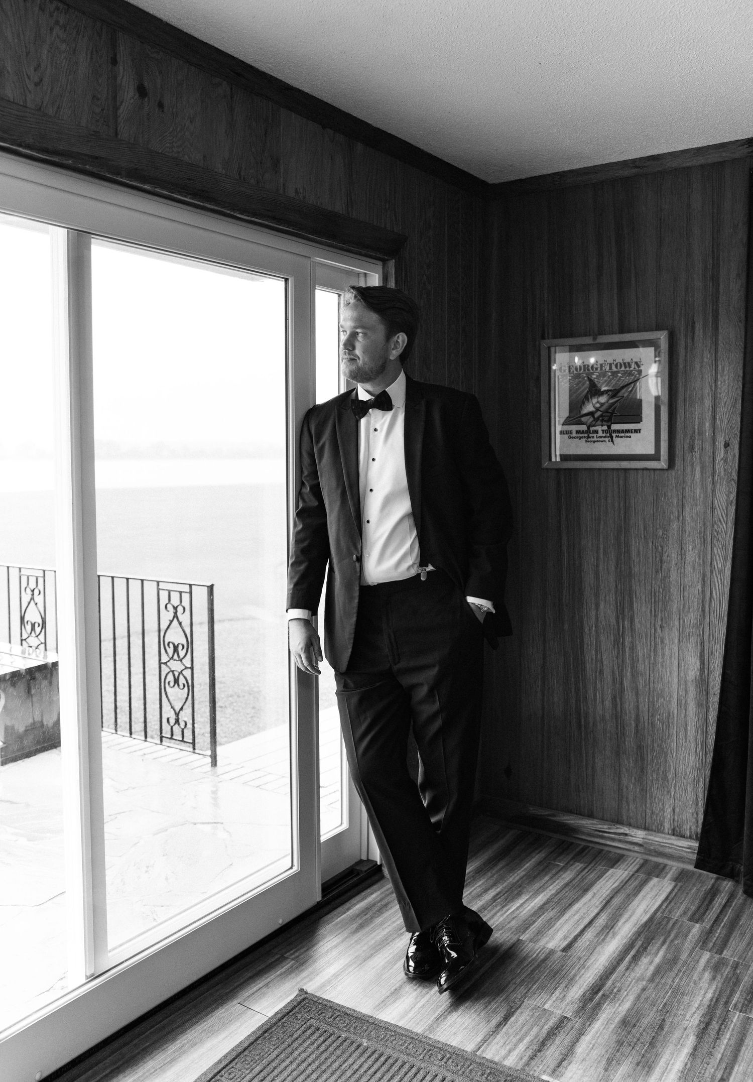 Groom in black tuxedo and bow tie standing by sliding glass door with ocean view in black and white photo.