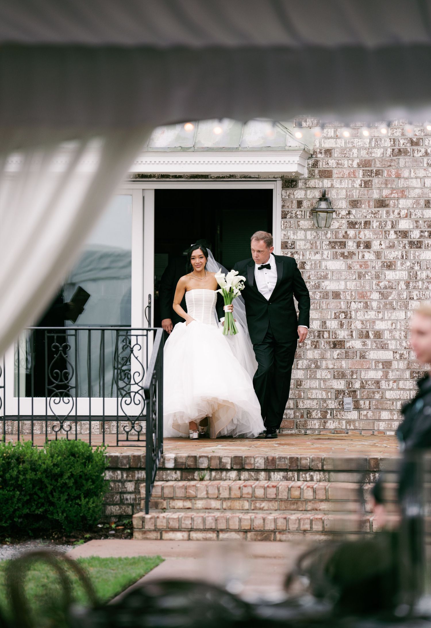 Bride in strapless ballgown holding calla lilies descends brick steps with escort through venue doorway.