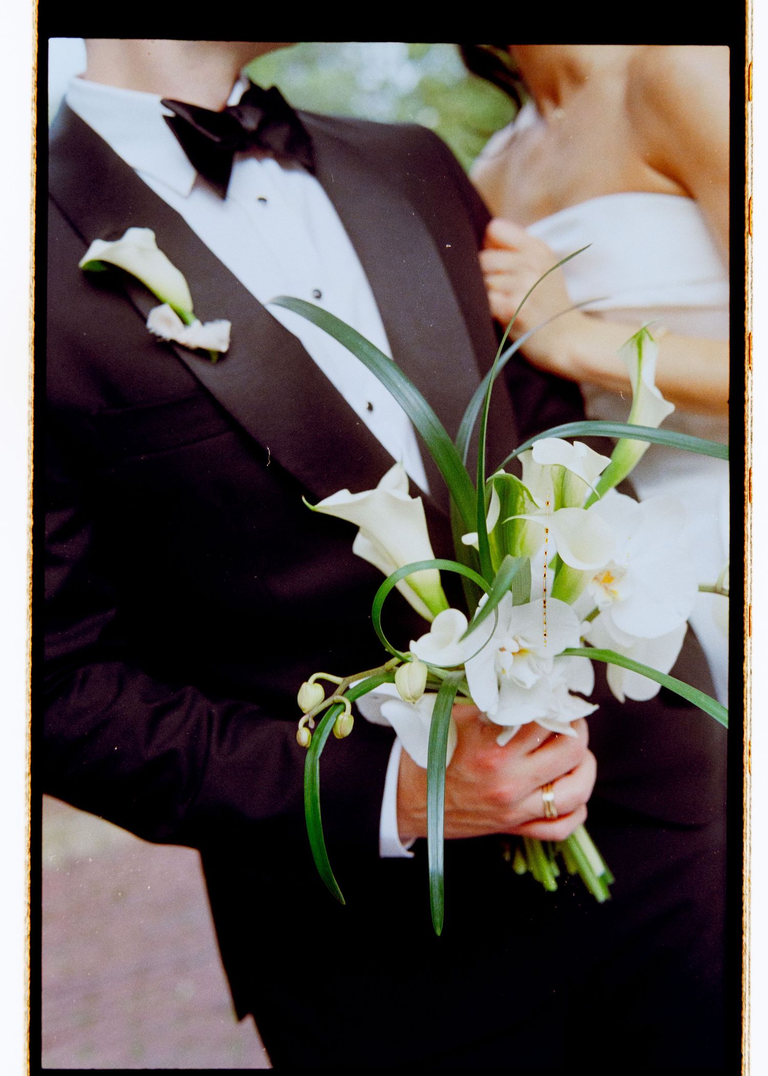 Groom in black tuxedo holding white calla lily bridal bouquet with green foliage accents.