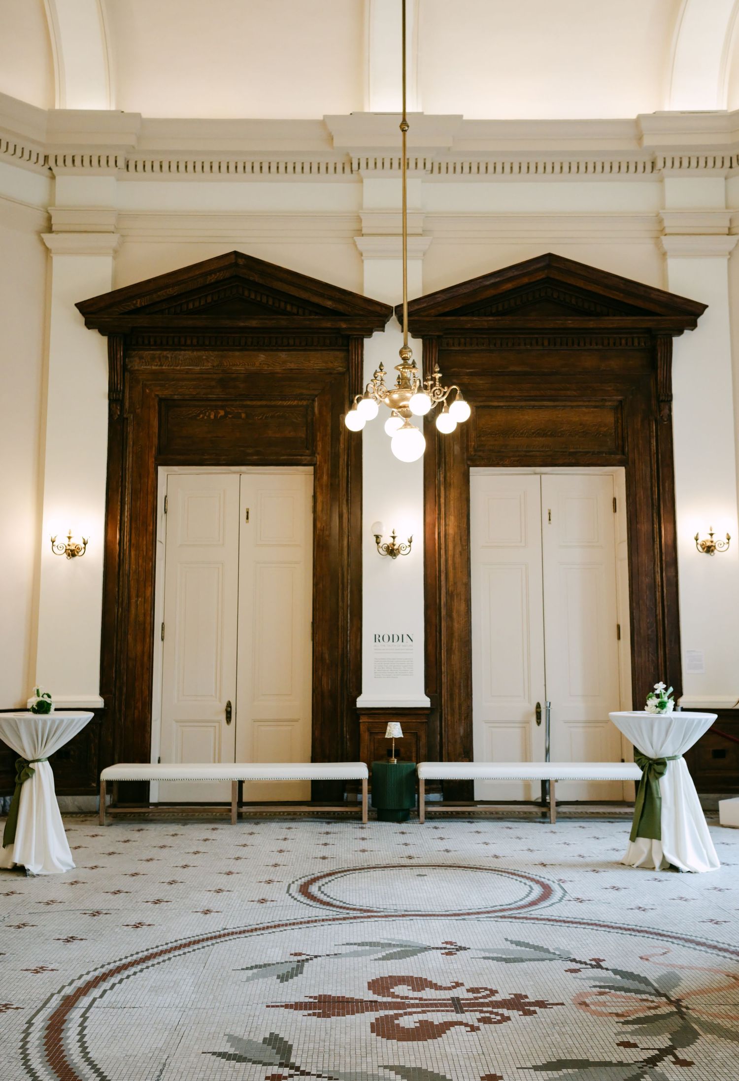 Grand wedding venue interior with mosaic floors, chandelier, dark wood doors, and draped cocktail tables.