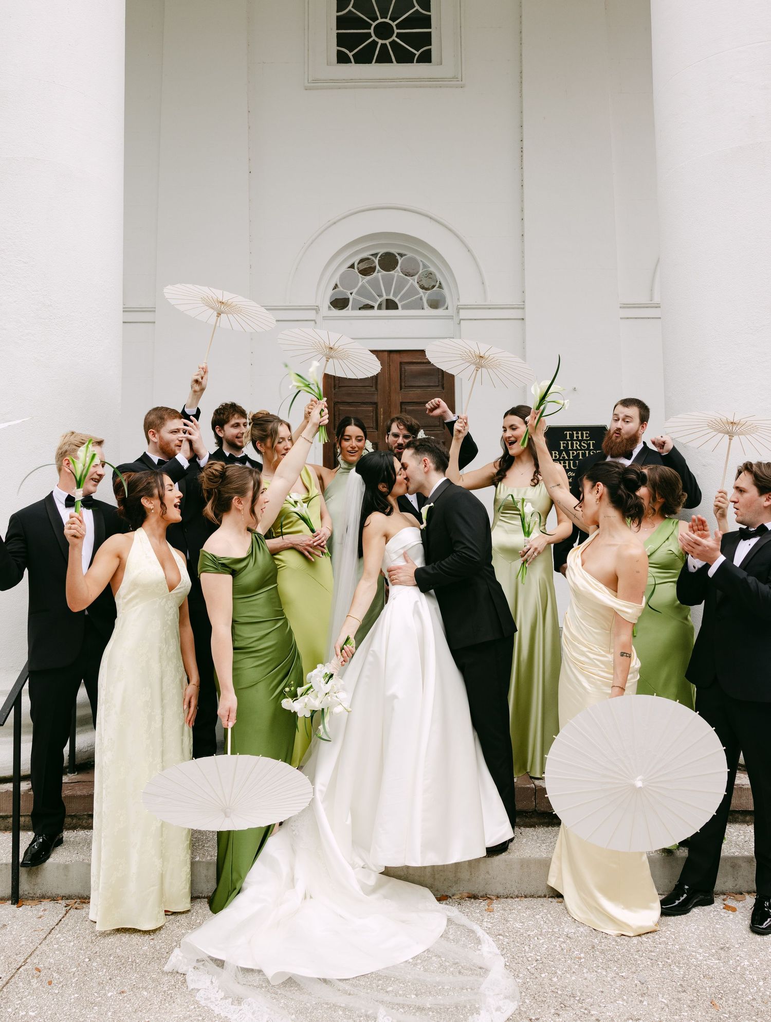 Wedding party celebrating on church steps, bridesmaids in green dresses holding white parasols with kissing bride and groom.