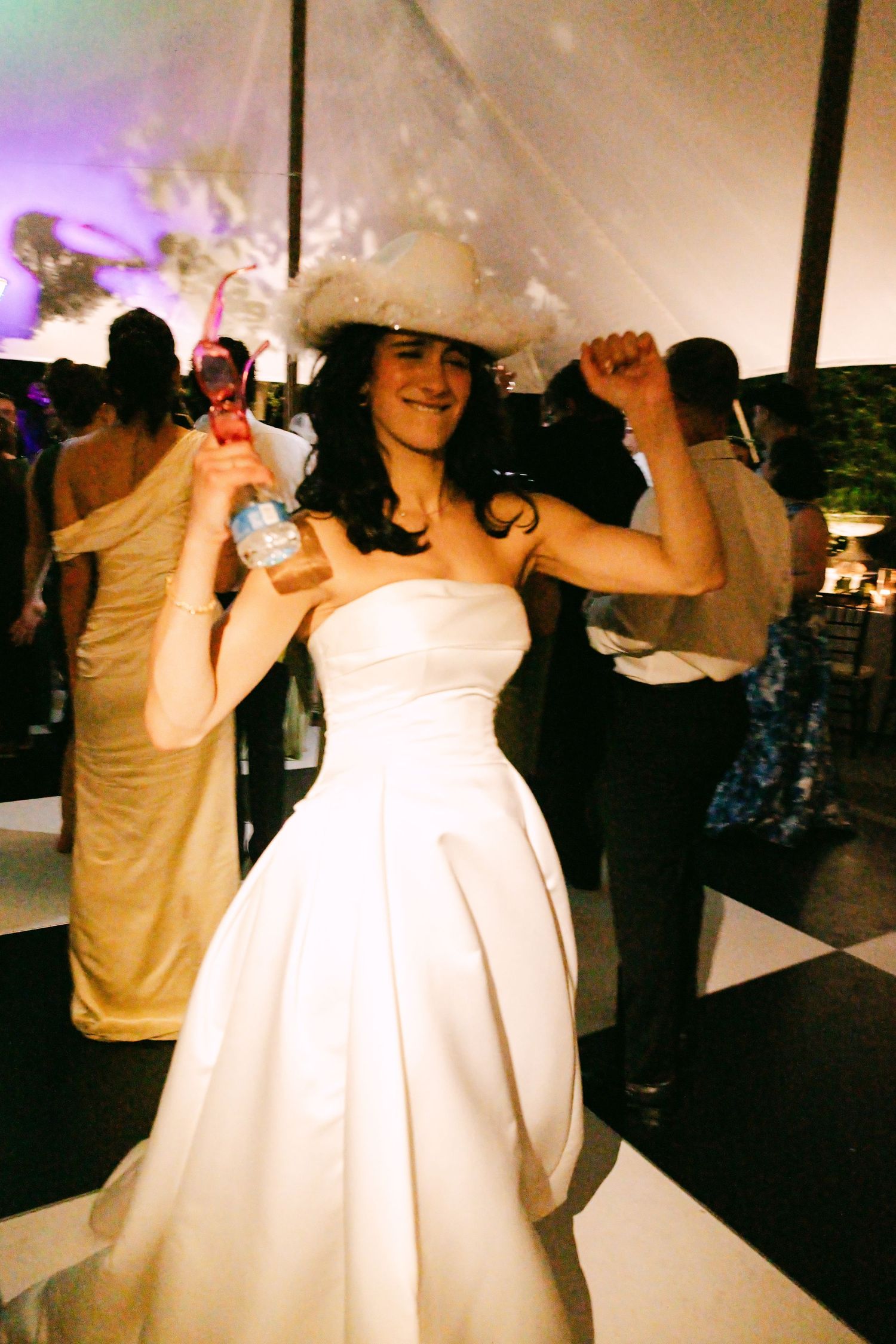 Bride in white strapless gown and white hat dances joyfully at wedding reception.