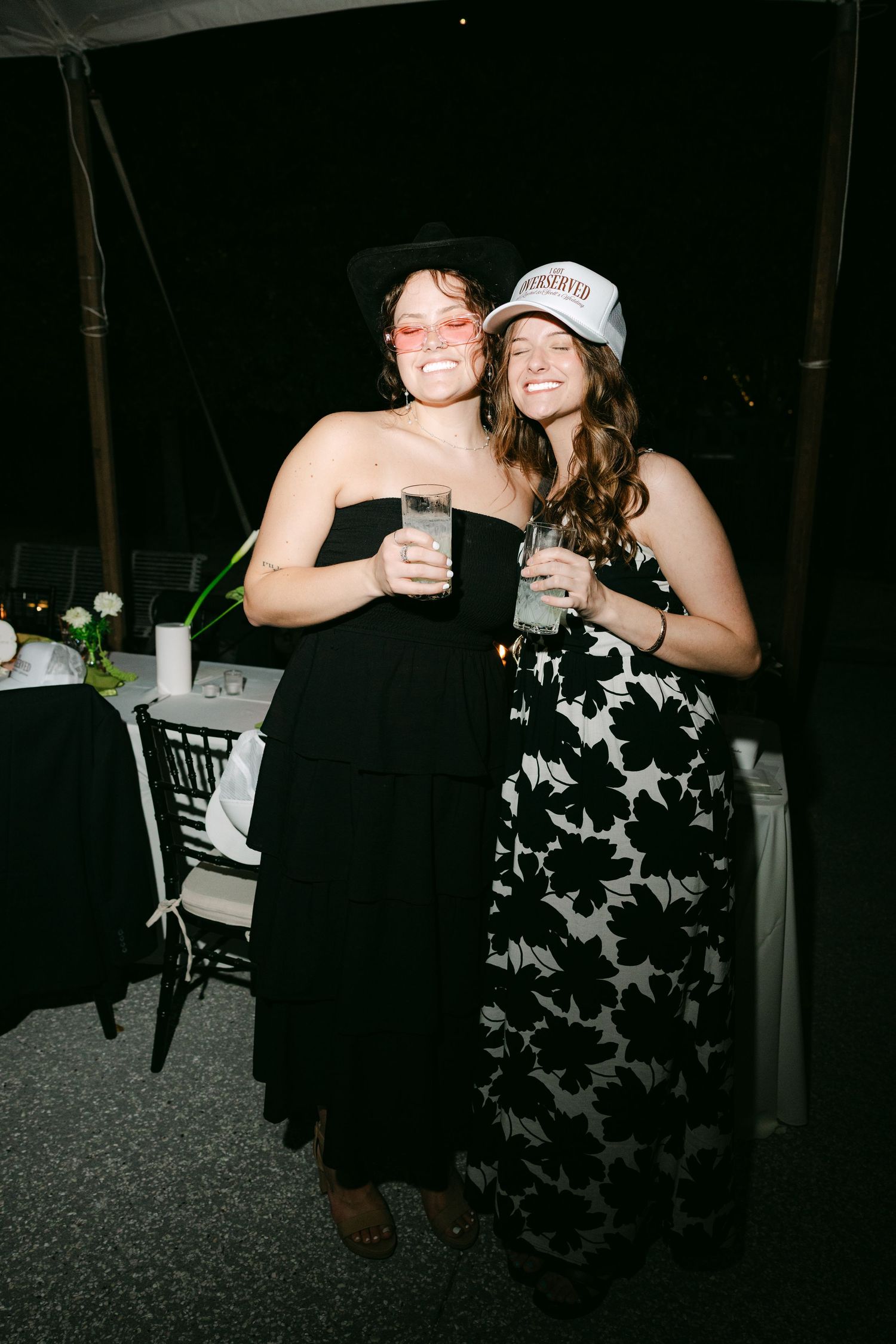 Two wedding guests smile and pose together wearing fun accessories at nighttime outdoor tented reception.