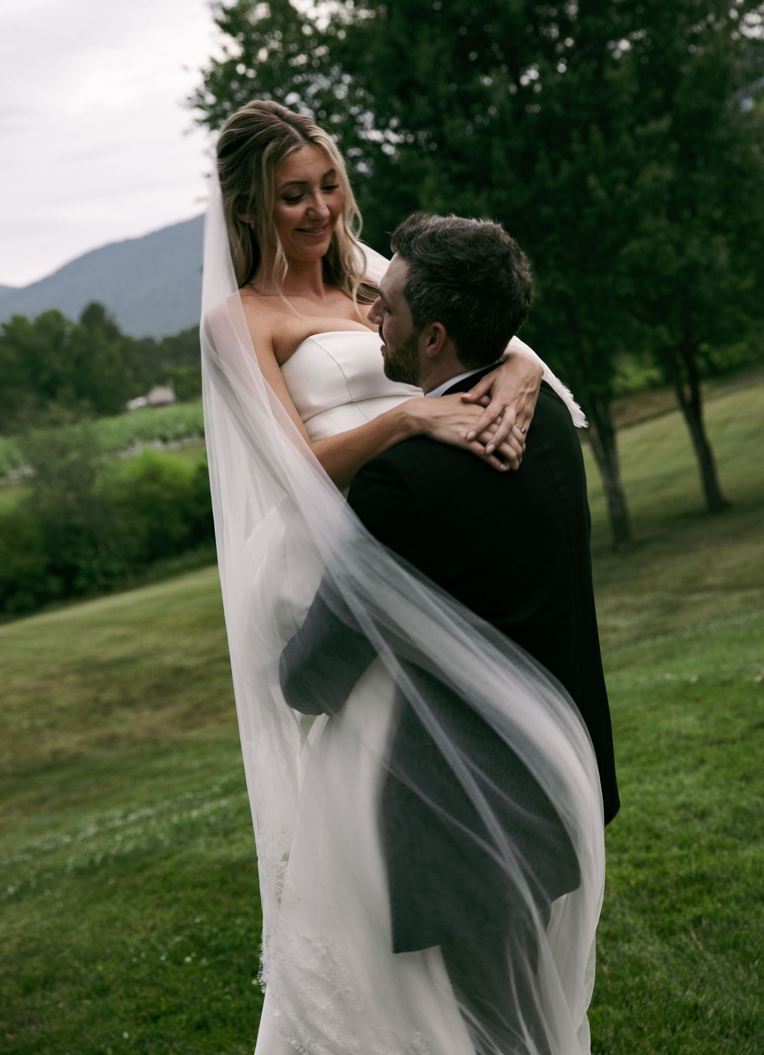 Groom lifting bride in white strapless gown and flowing veil outdoors with mountain backdrop.