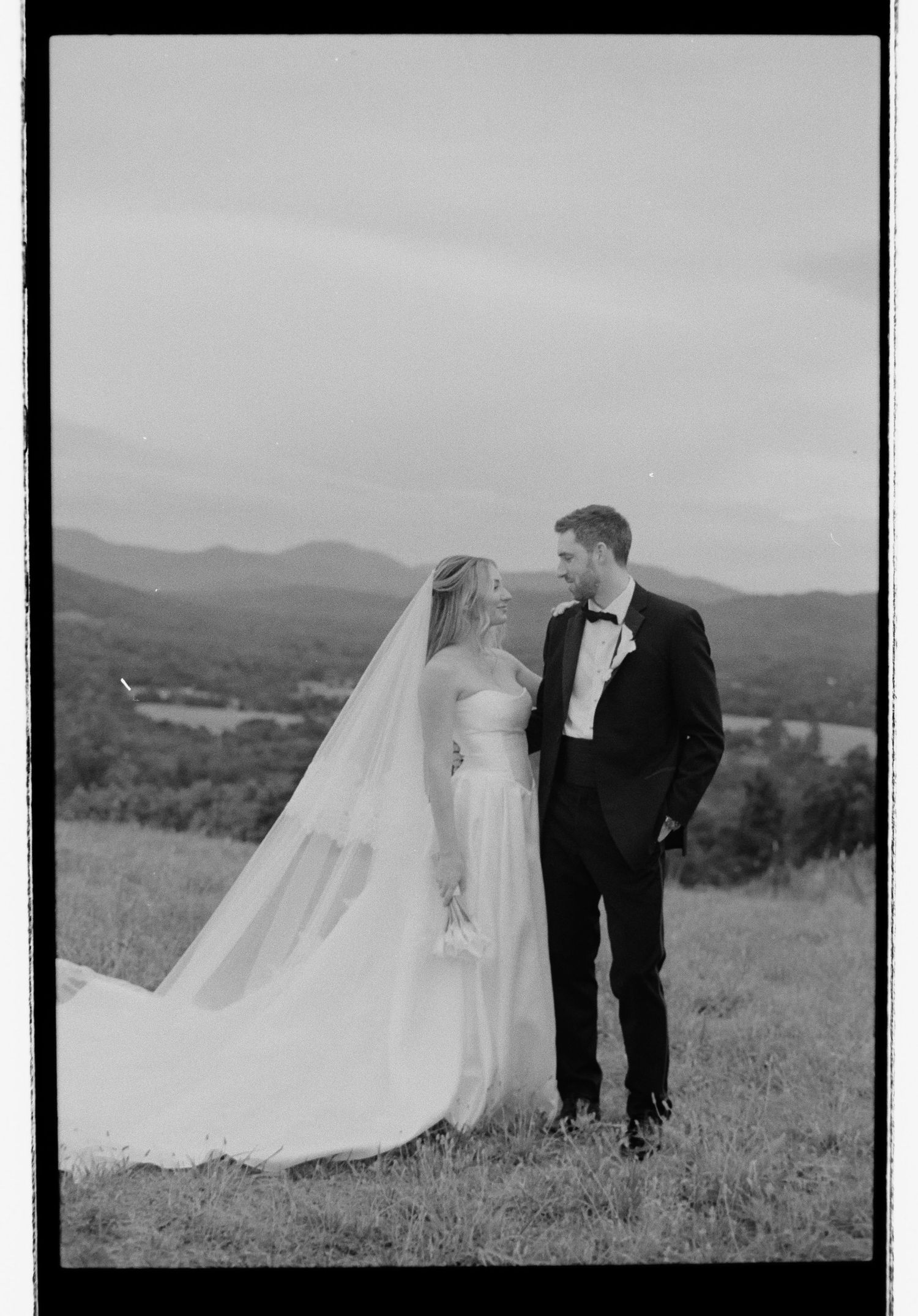 Black and white film photo of bride and groom sharing a moment on hilltop with stunning mountain and lake backdrop.