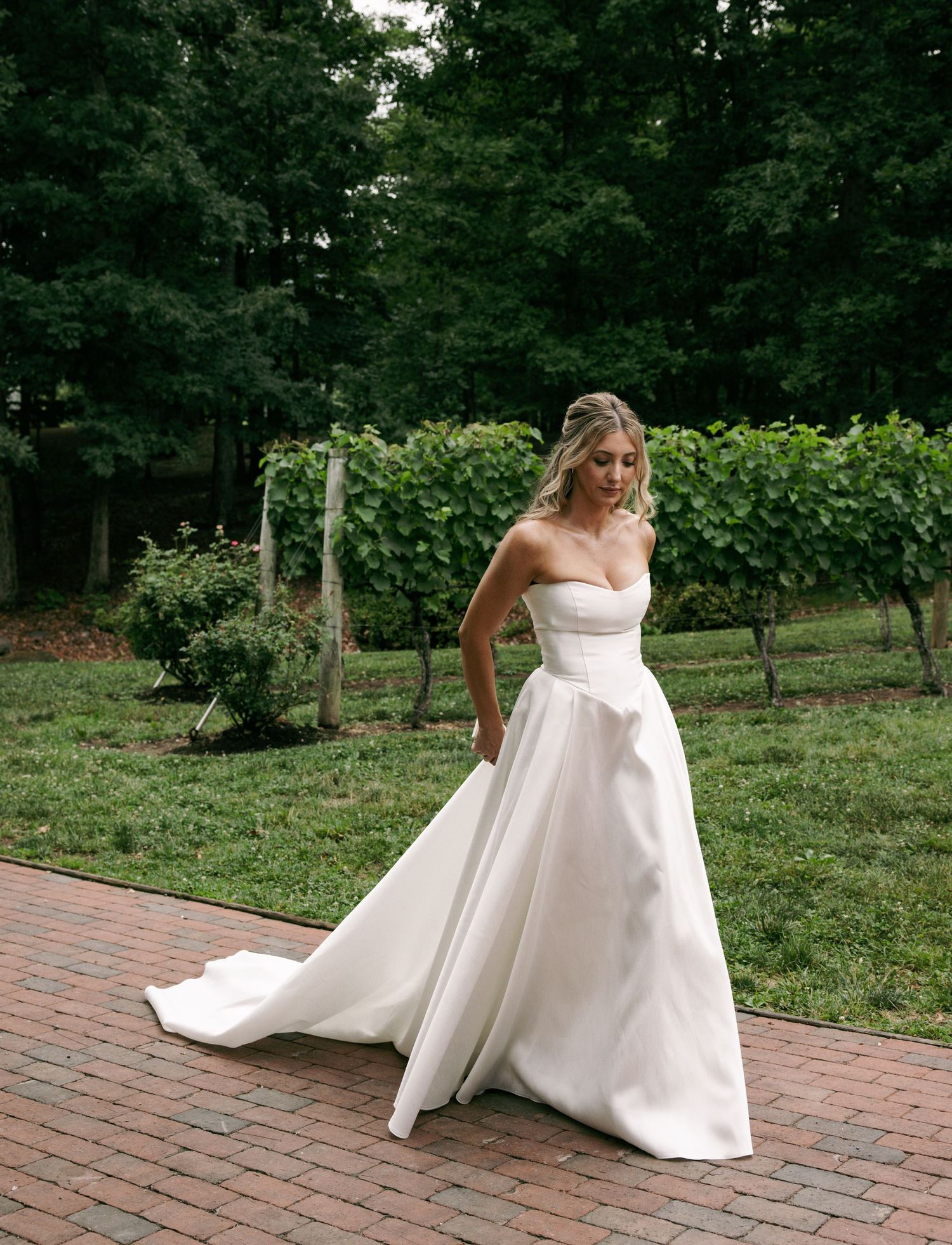 Bride in strapless white satin ball gown with train poses on brick path surrounded by lush vineyard greenery.