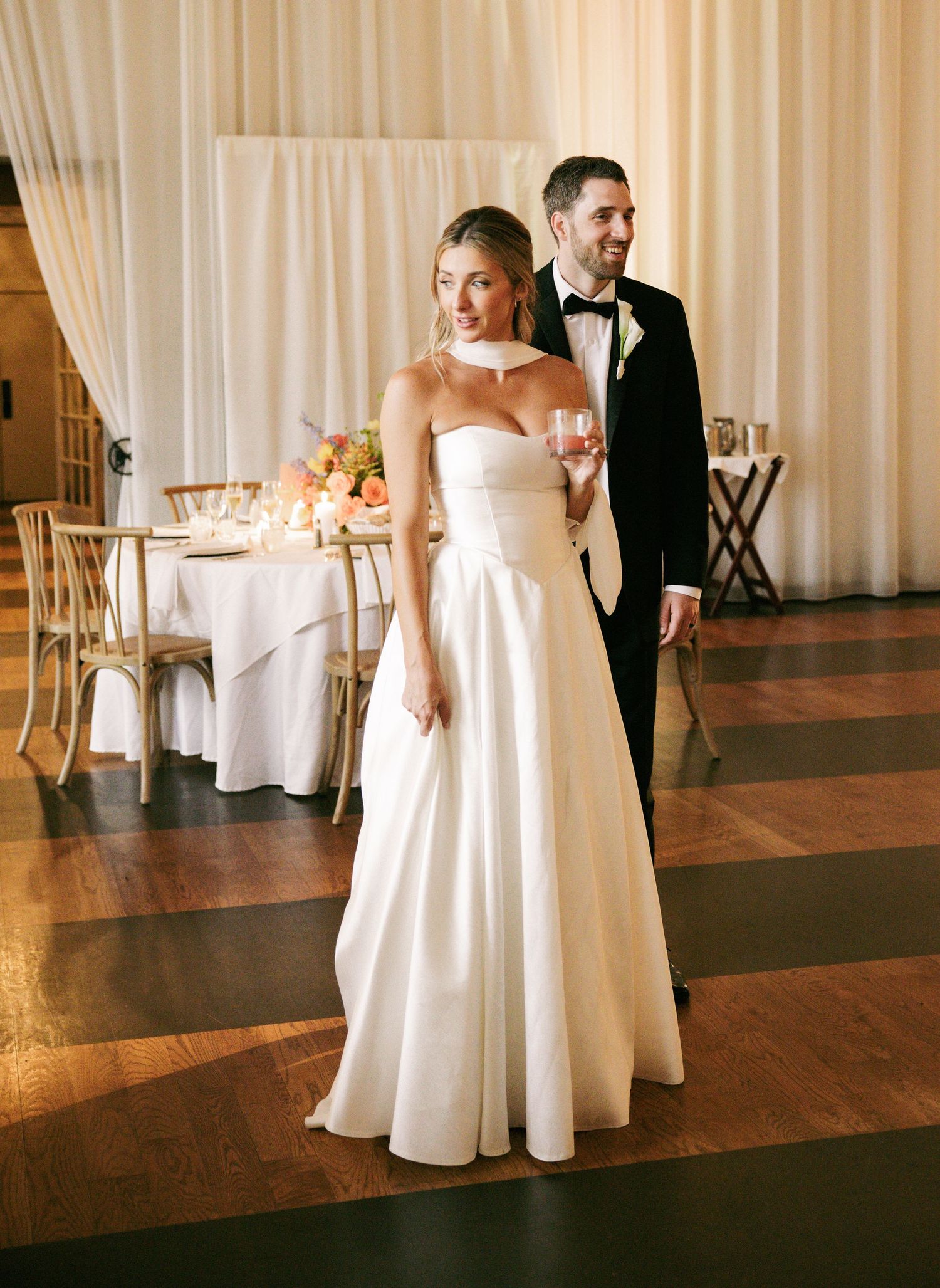 Bride in strapless white ball gown stands with groom in black tuxedo on wooden dance floor.