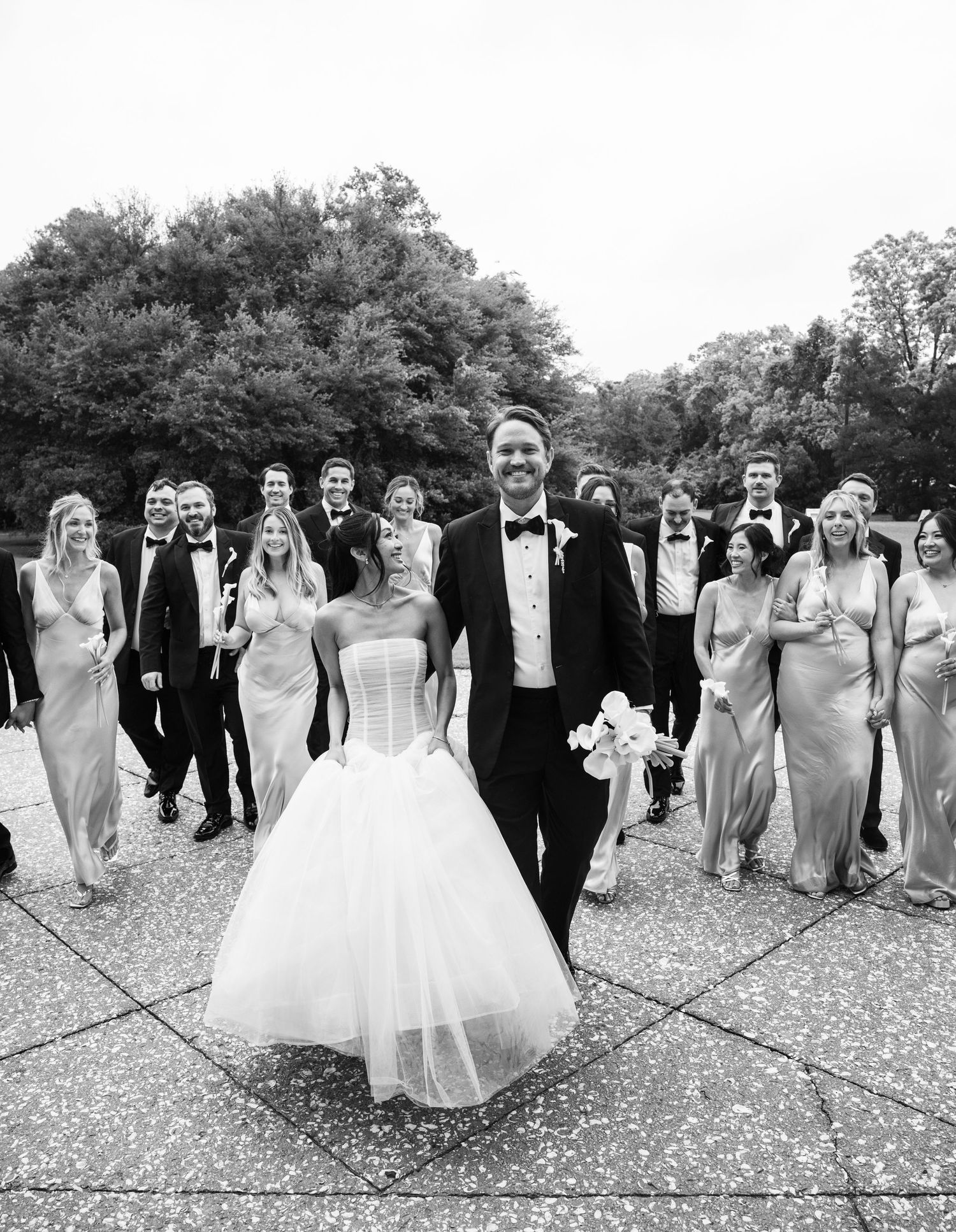 Black and white photo of bride and groom leading wedding party across outdoor plaza, trees in background.