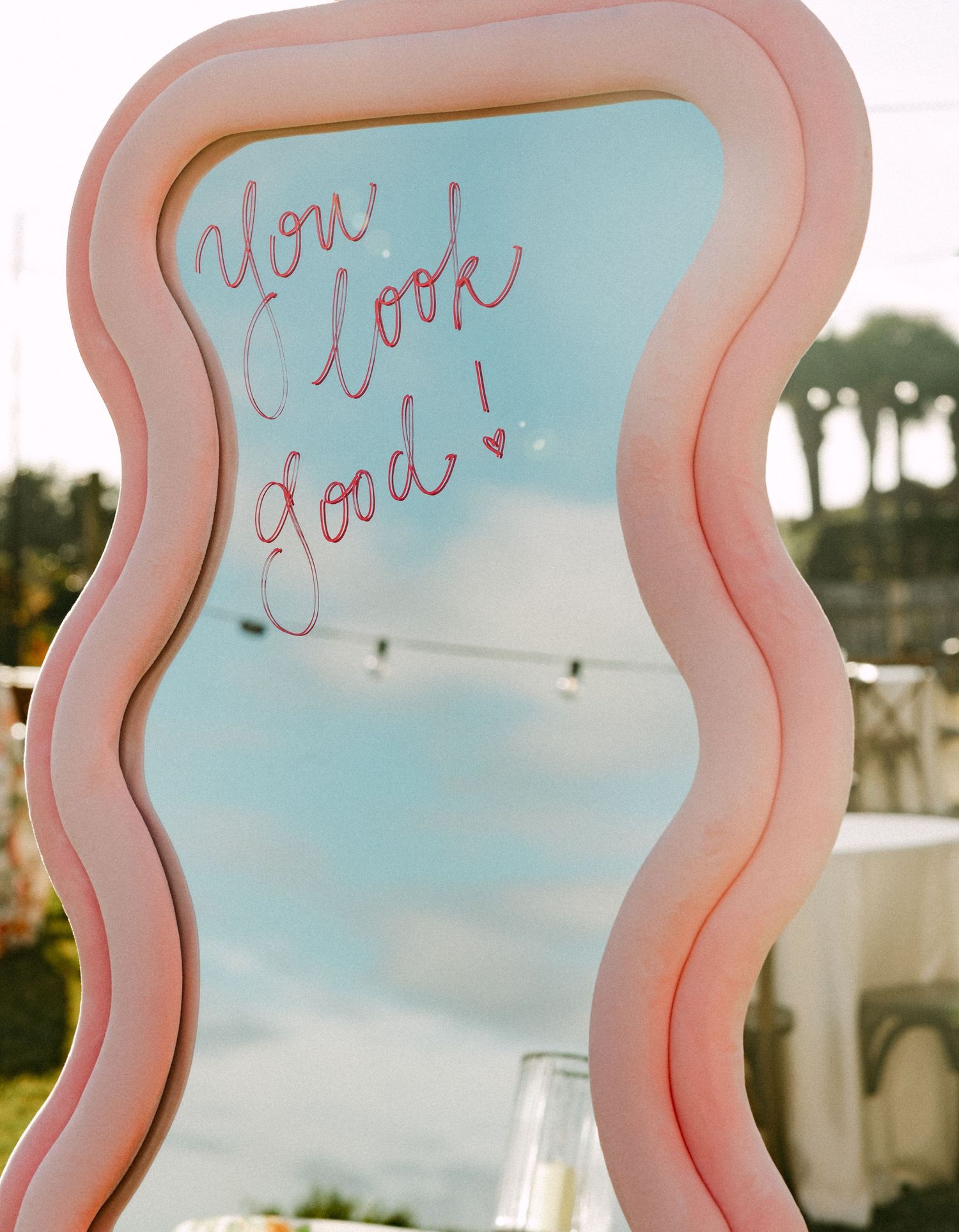 Decorative wavy pink mirror with You Look Good written in script at outdoor wedding.