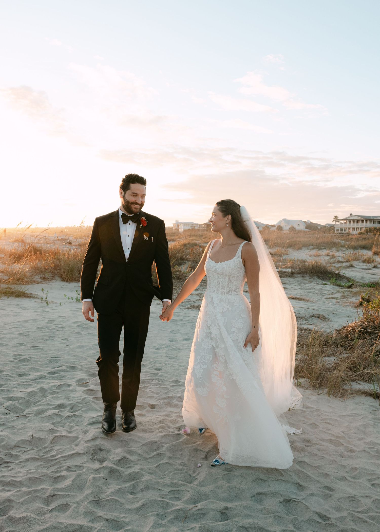 Bride and groom walking hand in hand on a sandy beach at sunset in wedding attire.