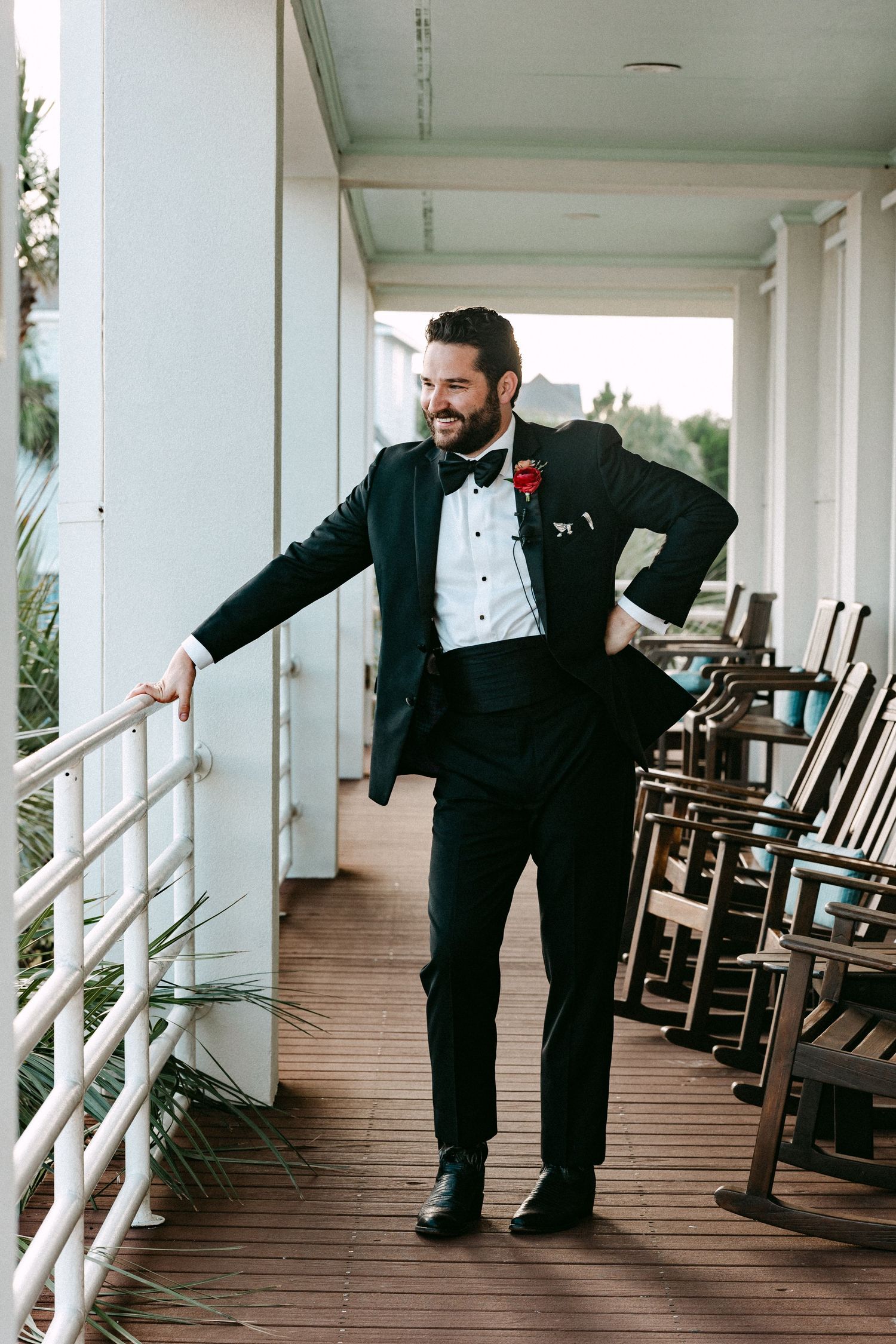 Smiling groom in black tuxedo with red rose boutonniere posing on a hotel porch.