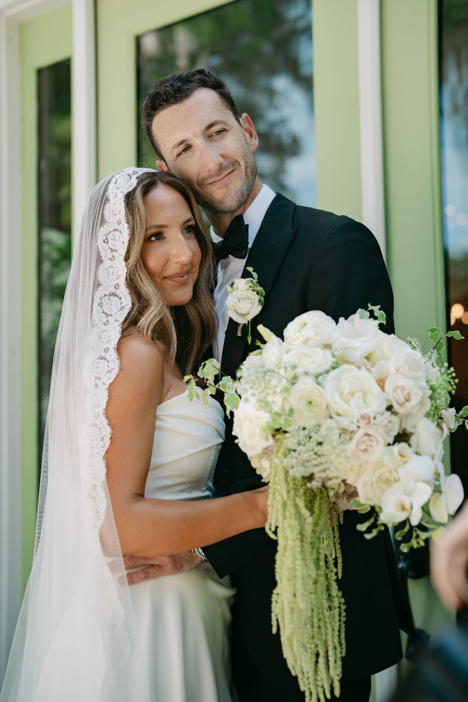 A romantic wedding couple poses together in front of a green building with a beautiful white floral bouquet.