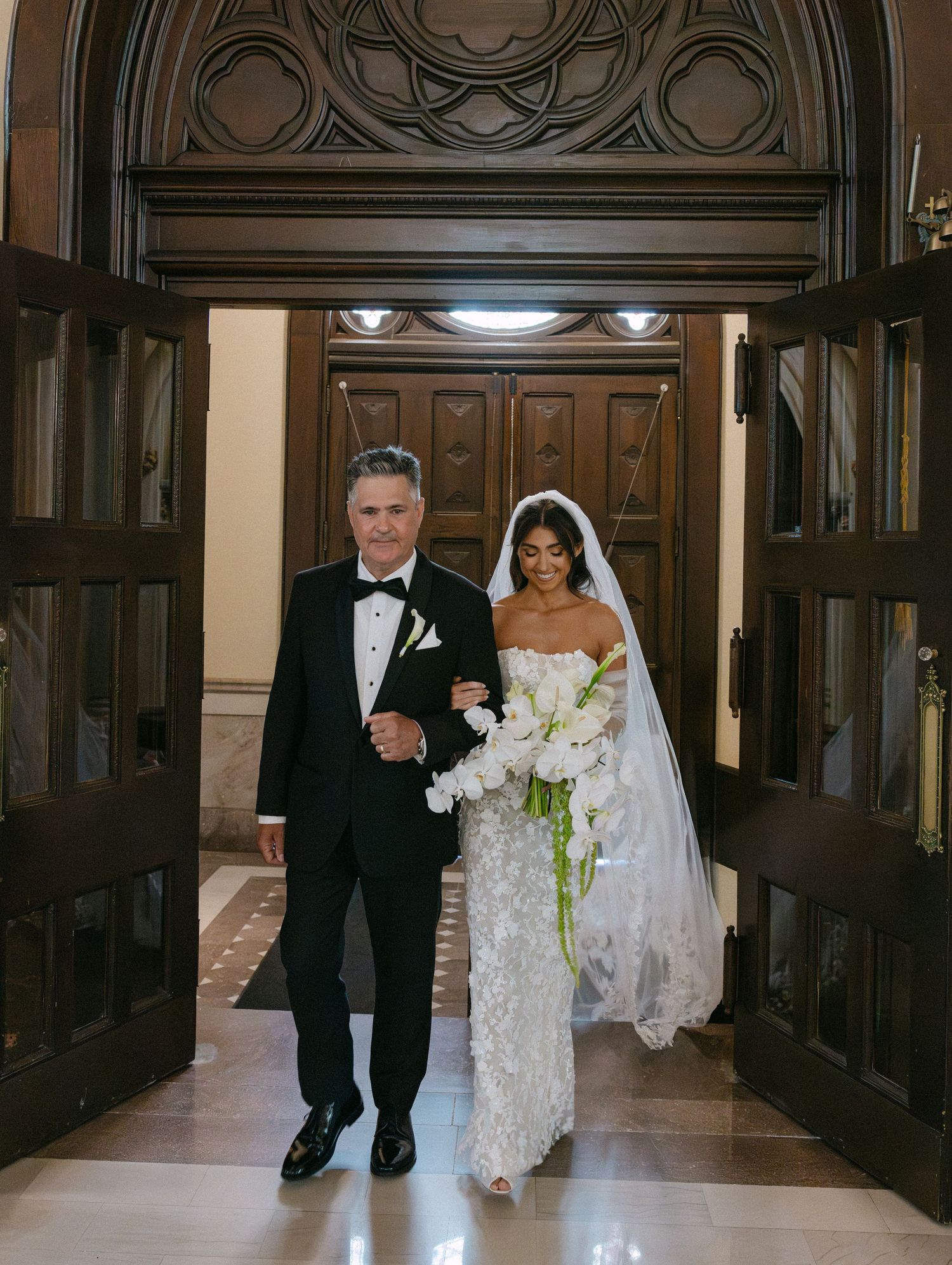 A traditional wedding procession walks down an elegant wood-paneled hallway.