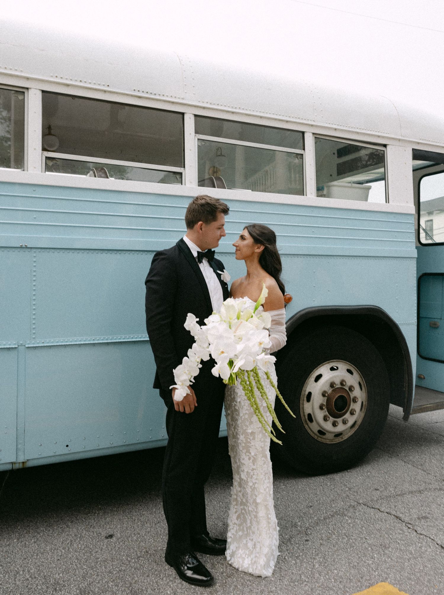 Newlyweds share intimate moment beside vintage blue bus with white orchid bouquet.