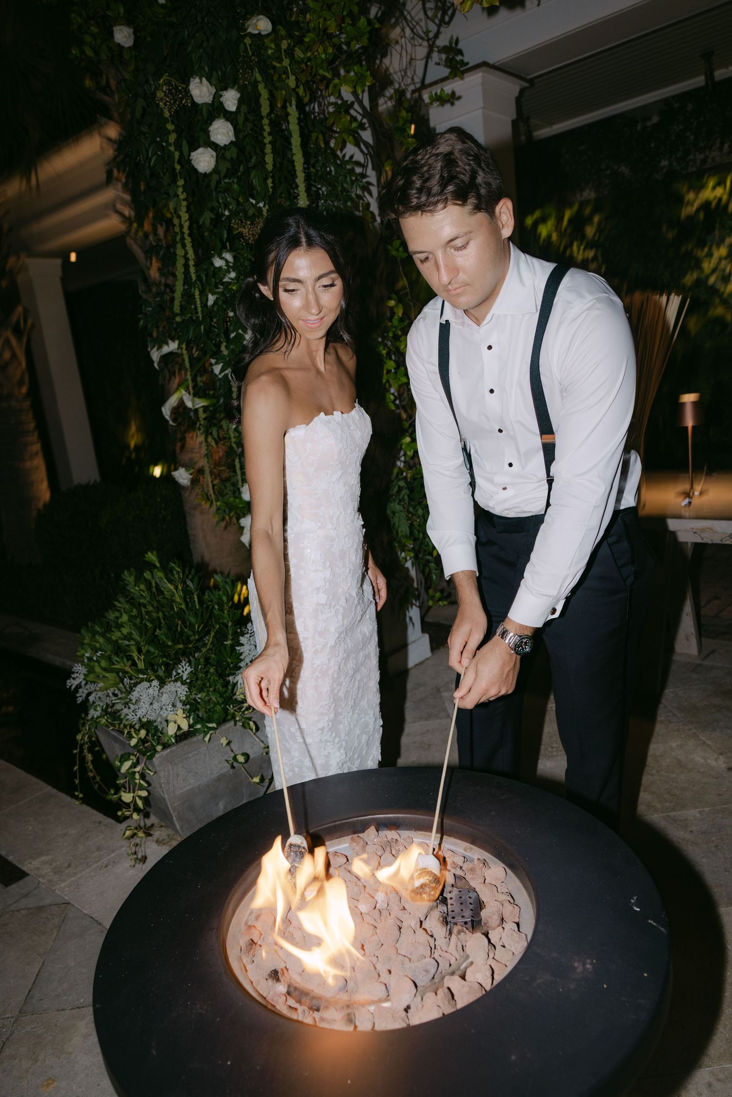 Couple roasting marshmallows at outdoor fire pit during wedding reception.