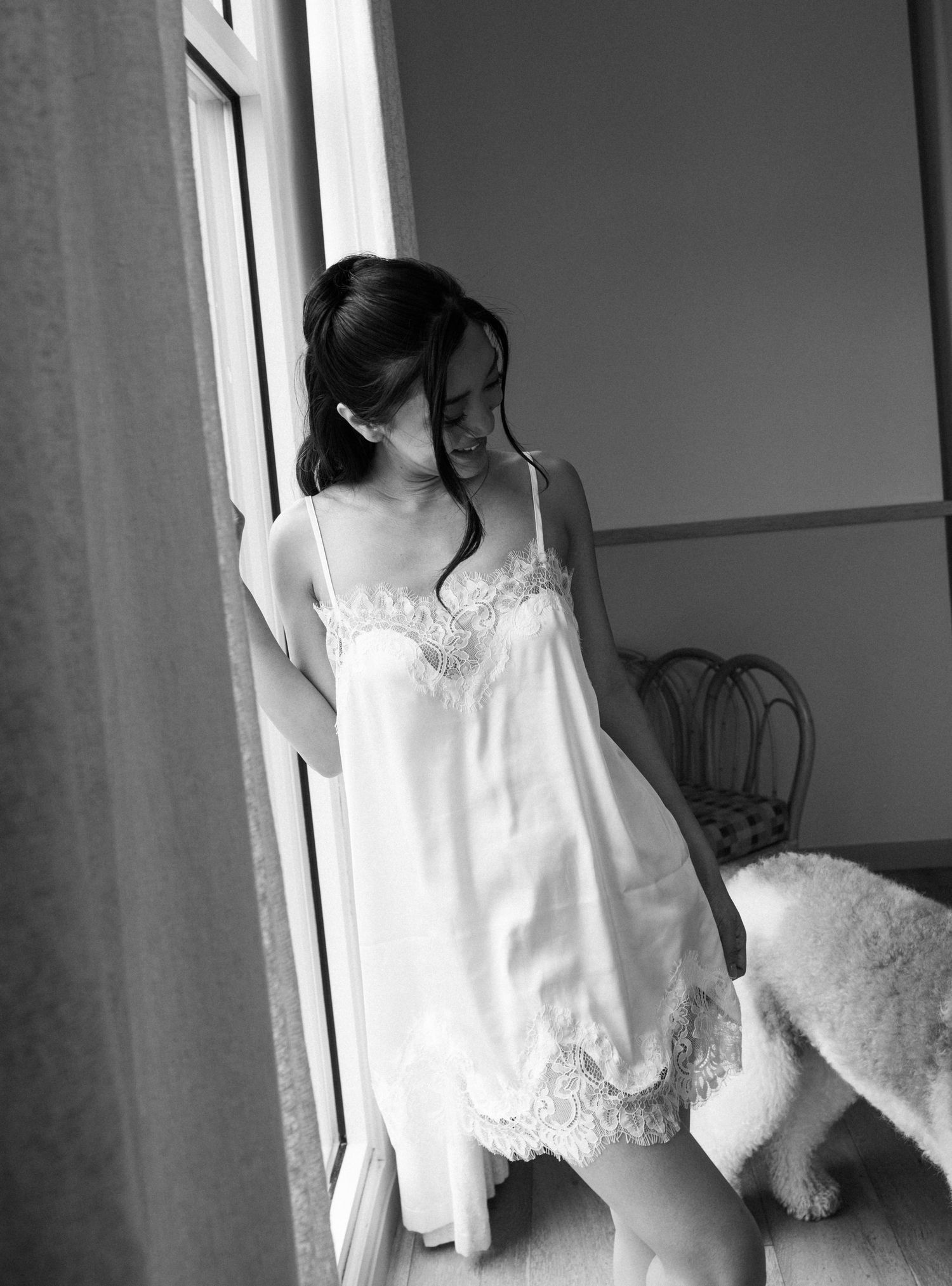 Black and white photo of a young woman in a white lace slip dress standing by a sunlit window.