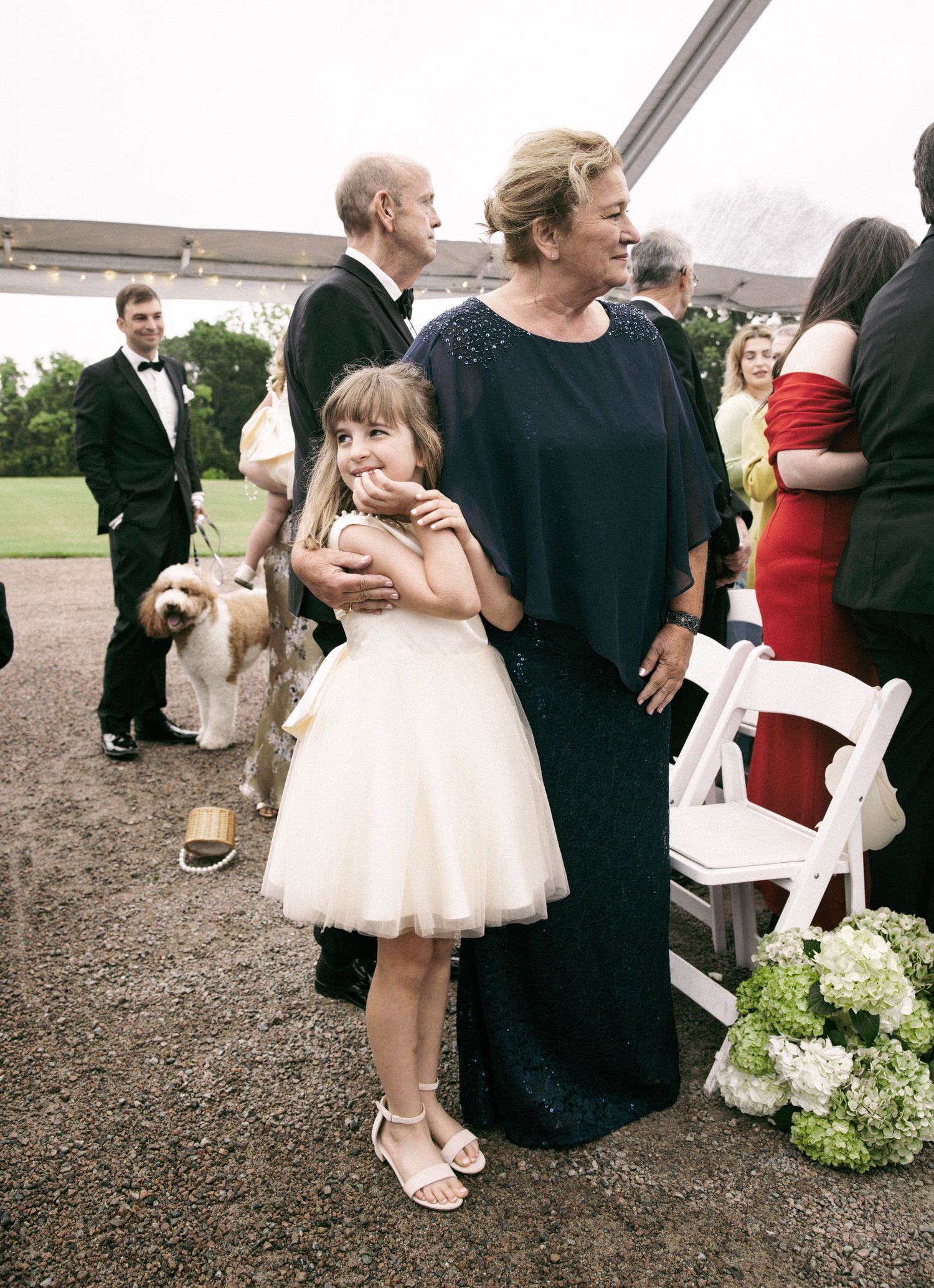 Grandmother in navy dress hugging flower girl in white dress at outdoor wedding.