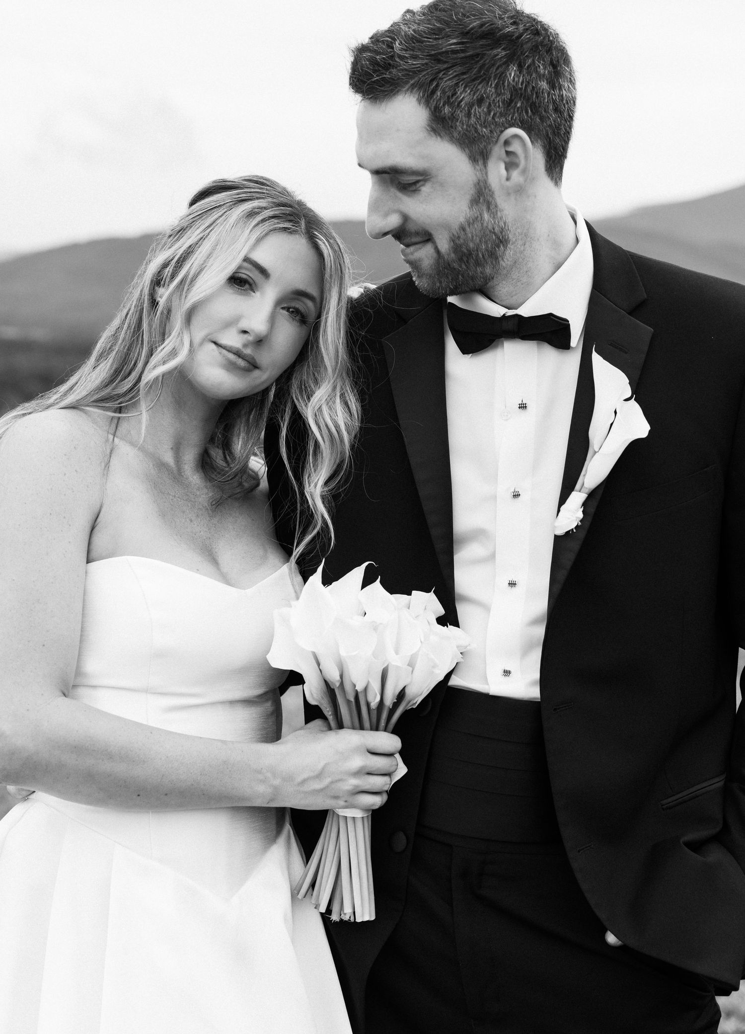 Black and white close-up of smiling bride holding white calla lily bouquet beside her tuxedo-clad groom.