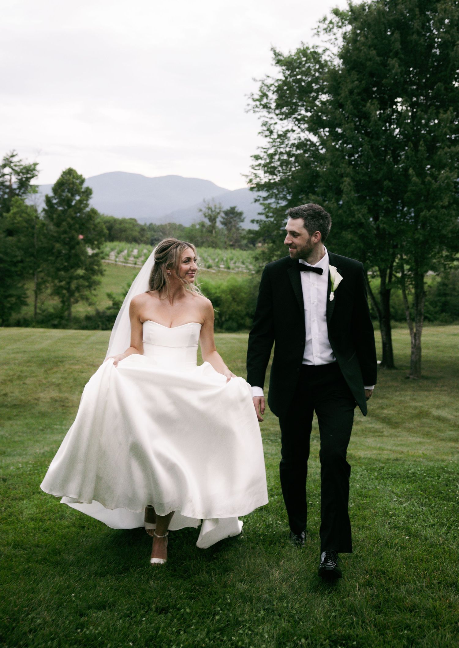 Bride in strapless white gown and groom in black tuxedo walking on green lawn with mountains.