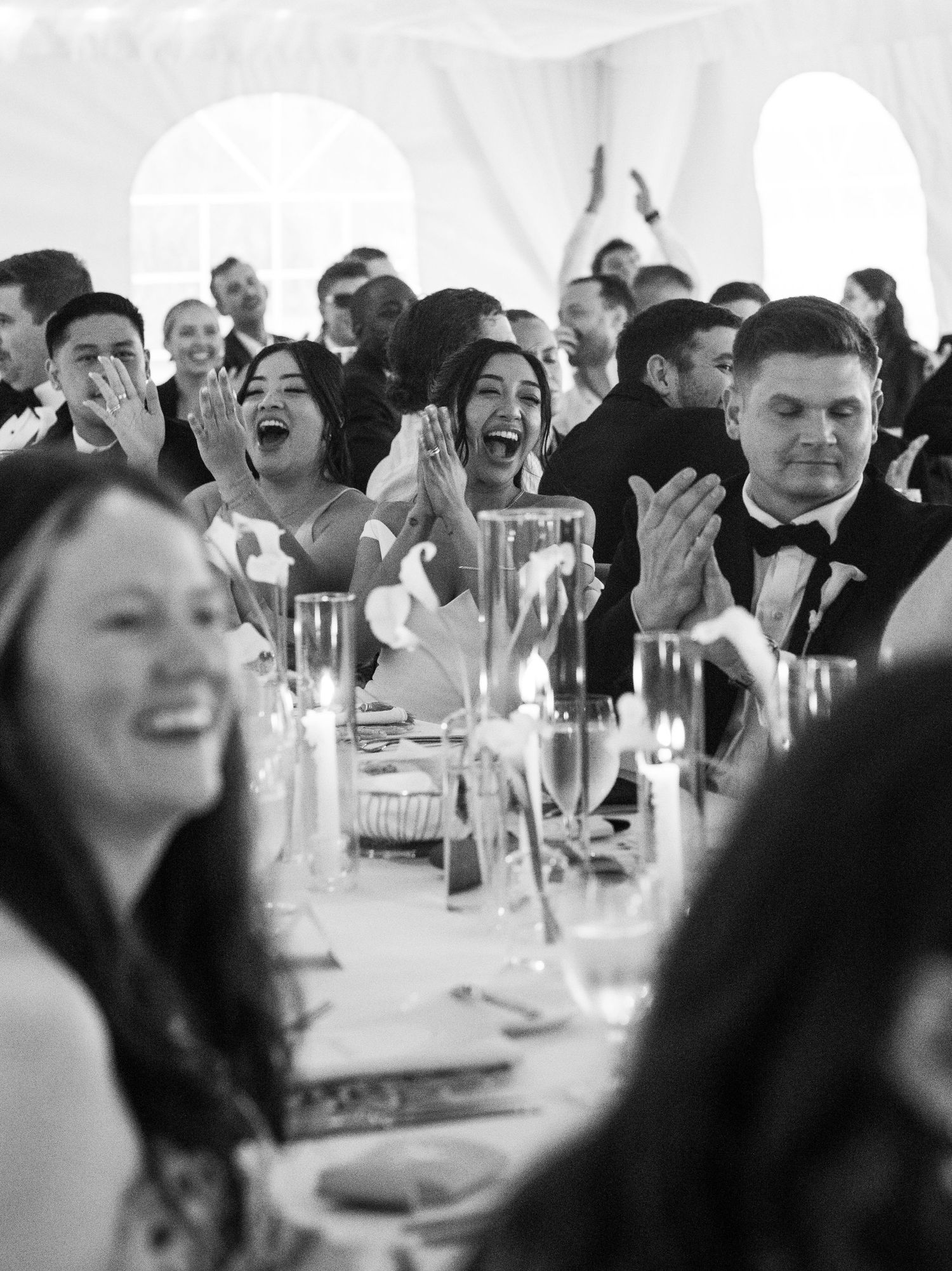 Black and white photo of joyful wedding guests laughing and clapping at a candlelit reception under a tent.