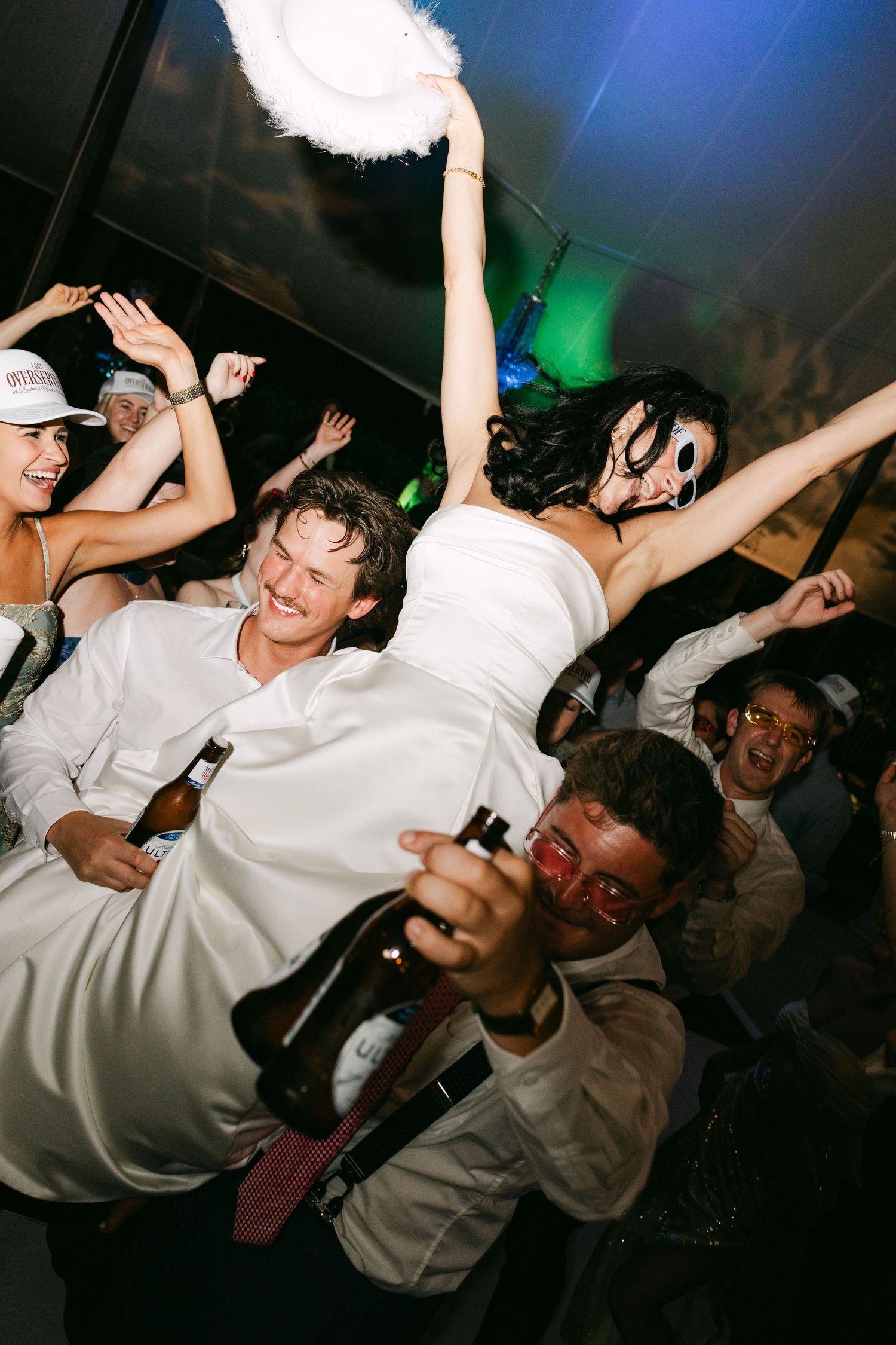 Bride in white gown wearing a mask being lifted by guests during an energetic wedding reception dance.