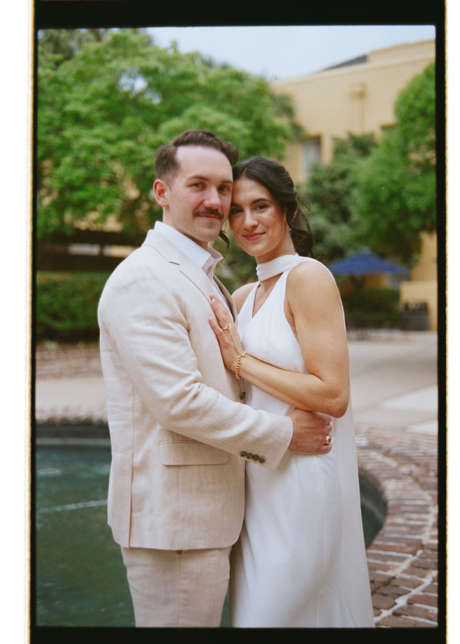 Couple poses lovingly in casual attire near a fountain in a film photography style portrait.