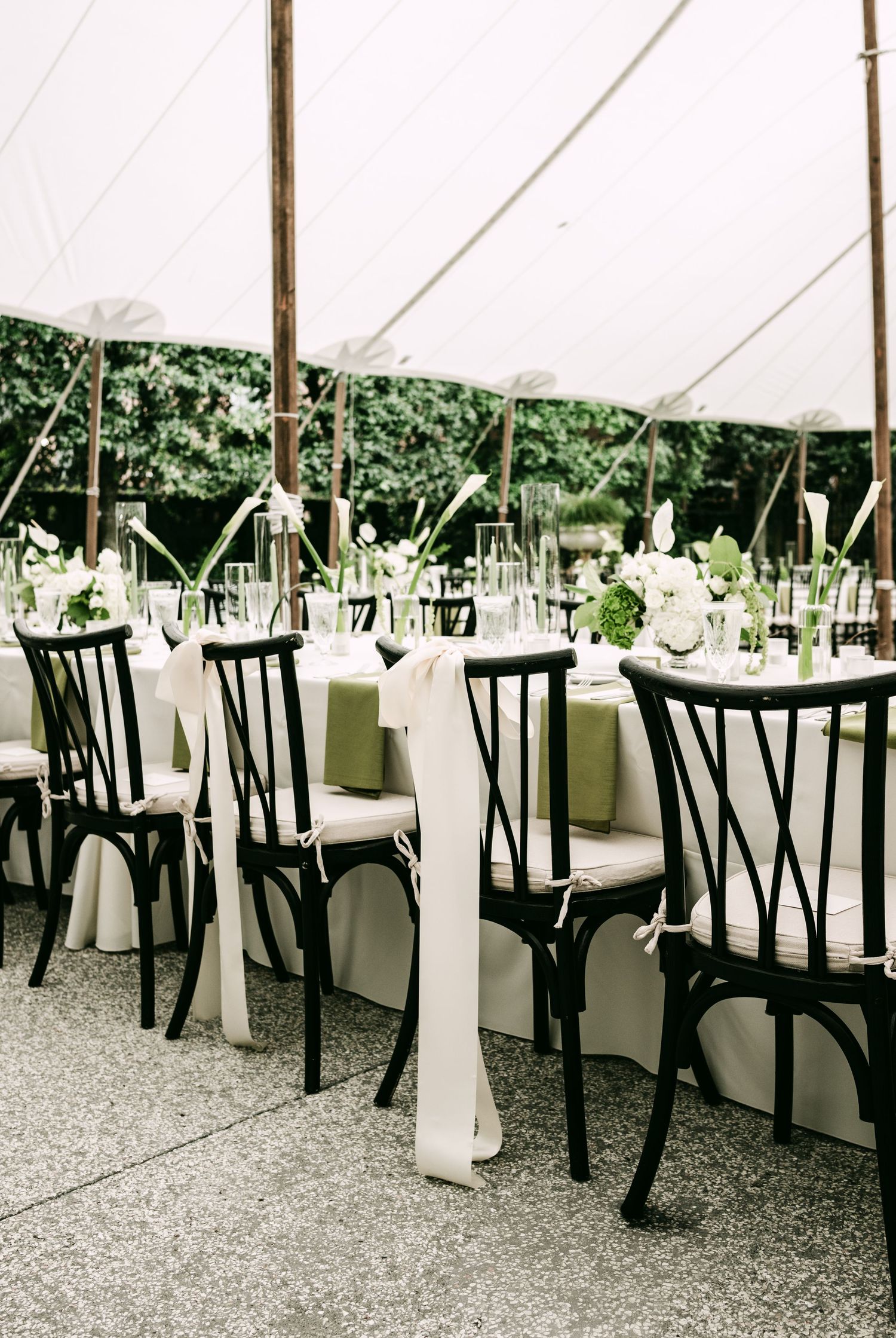 Elegant wedding reception tables with black cross-back chairs, white linens, and green floral centerpieces under a white tent.