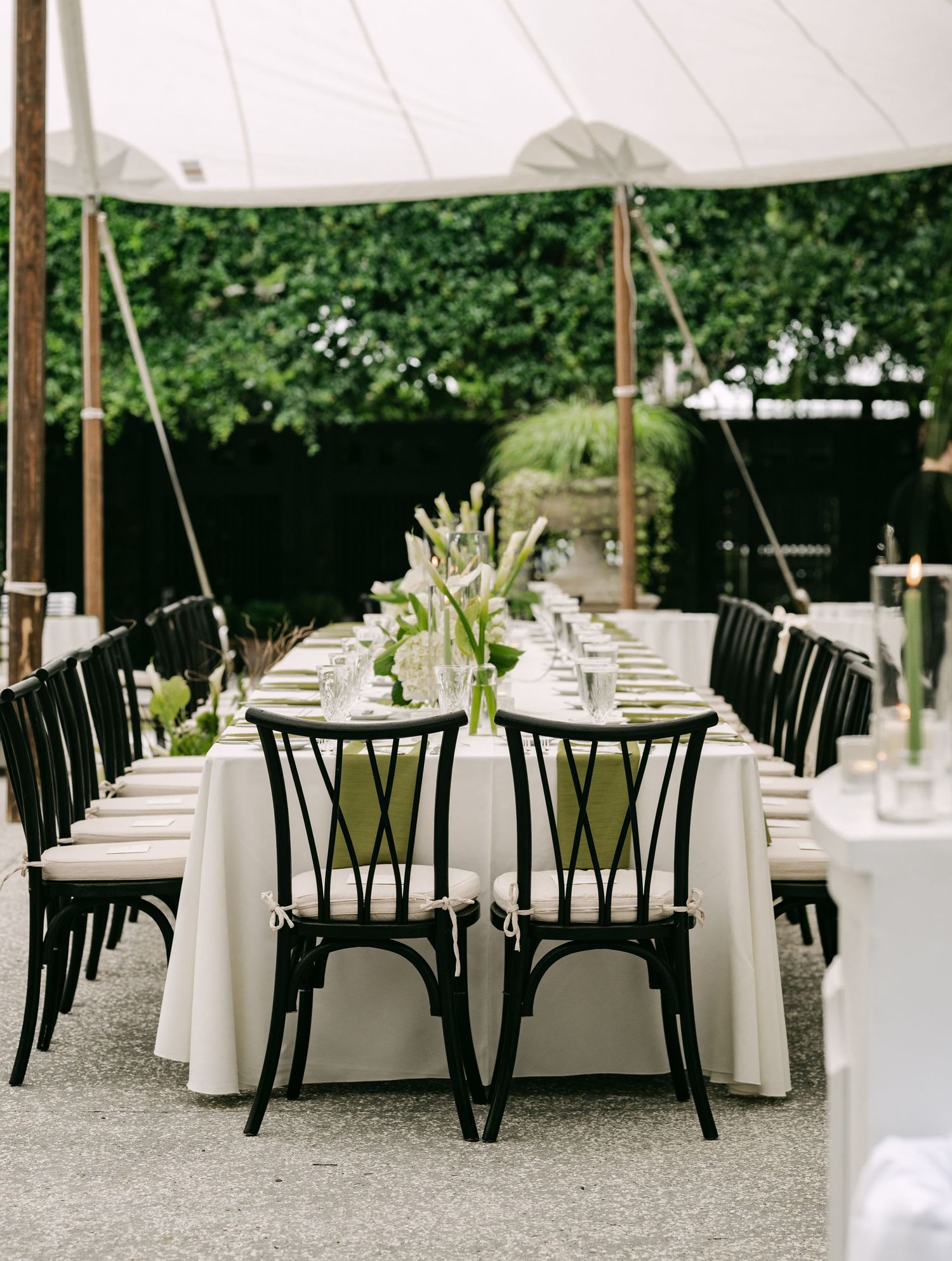 Outdoor wedding reception under white tent featuring black chairs, white tablecloths, and tall green floral arrangements.