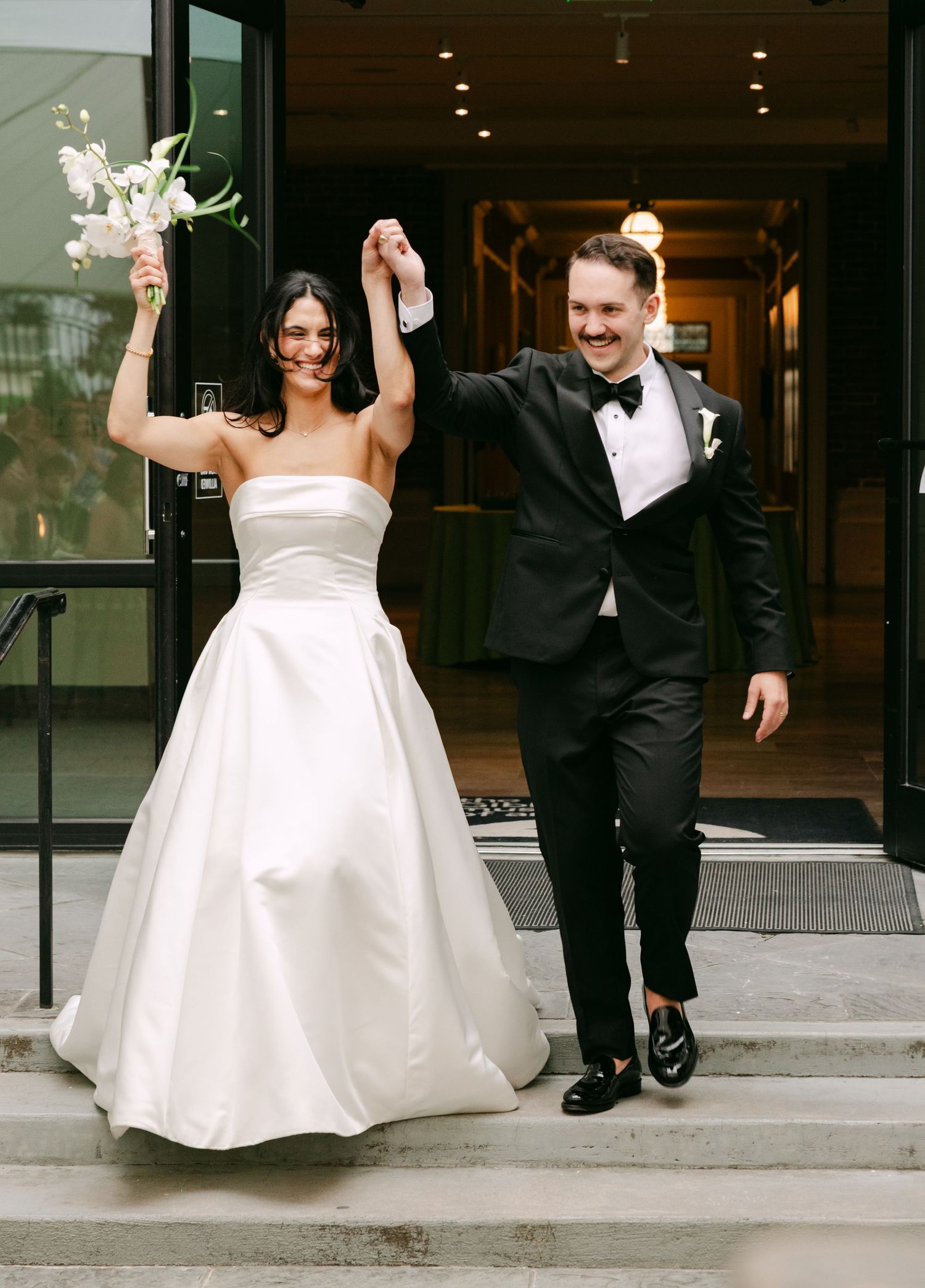 Joyful newlyweds celebrate on steps, bride in strapless ball gown holding bouquet, groom in black tuxedo raising hands.