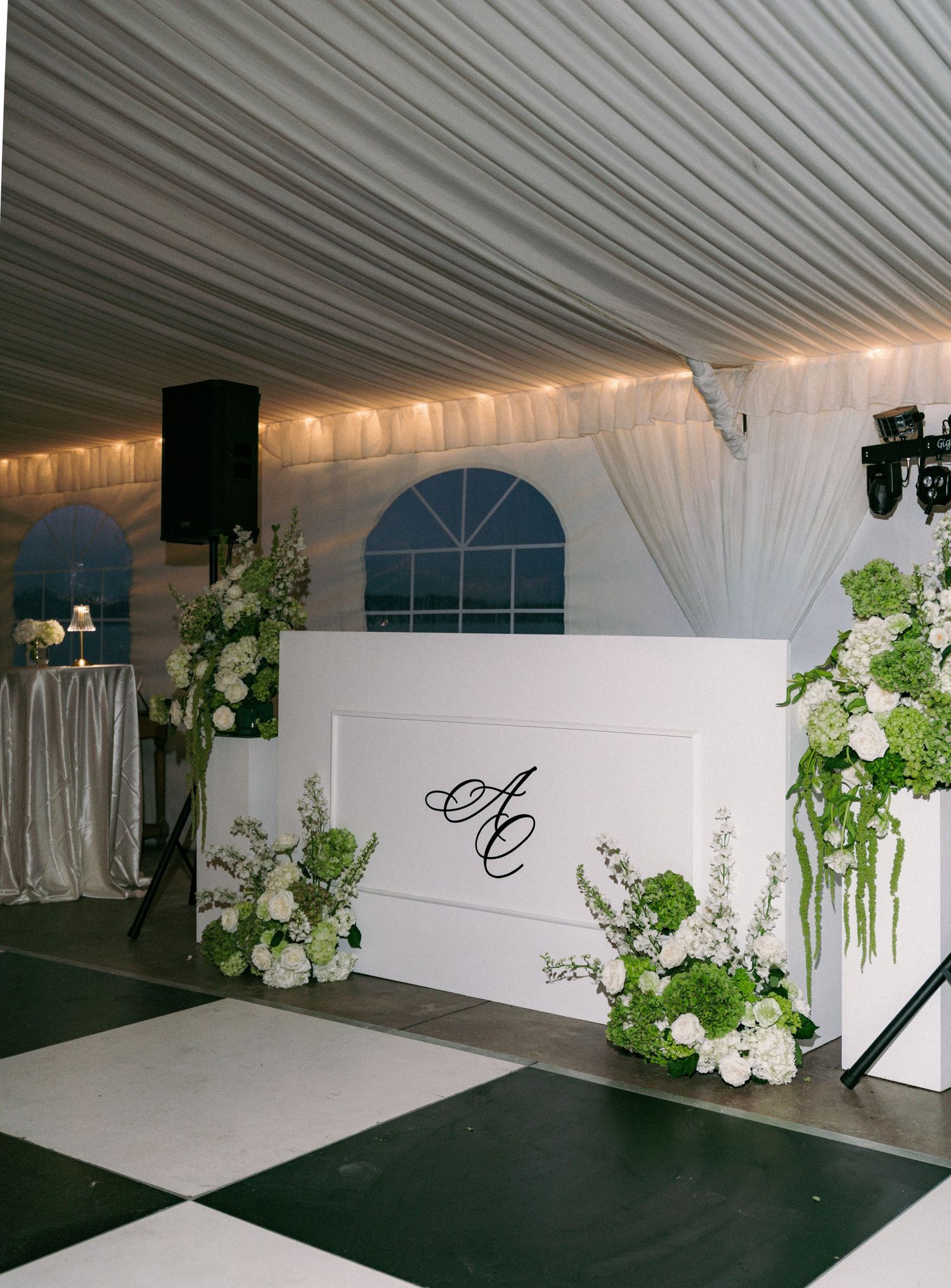 Elegant white wedding DJ booth with monogram AC and lush green and white floral arrangements.