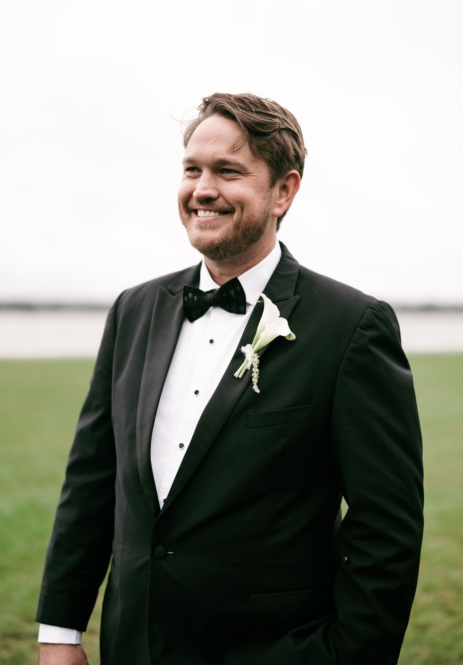 Handsome groom in black tuxedo with white boutonniere smiling outdoors on his wedding day.